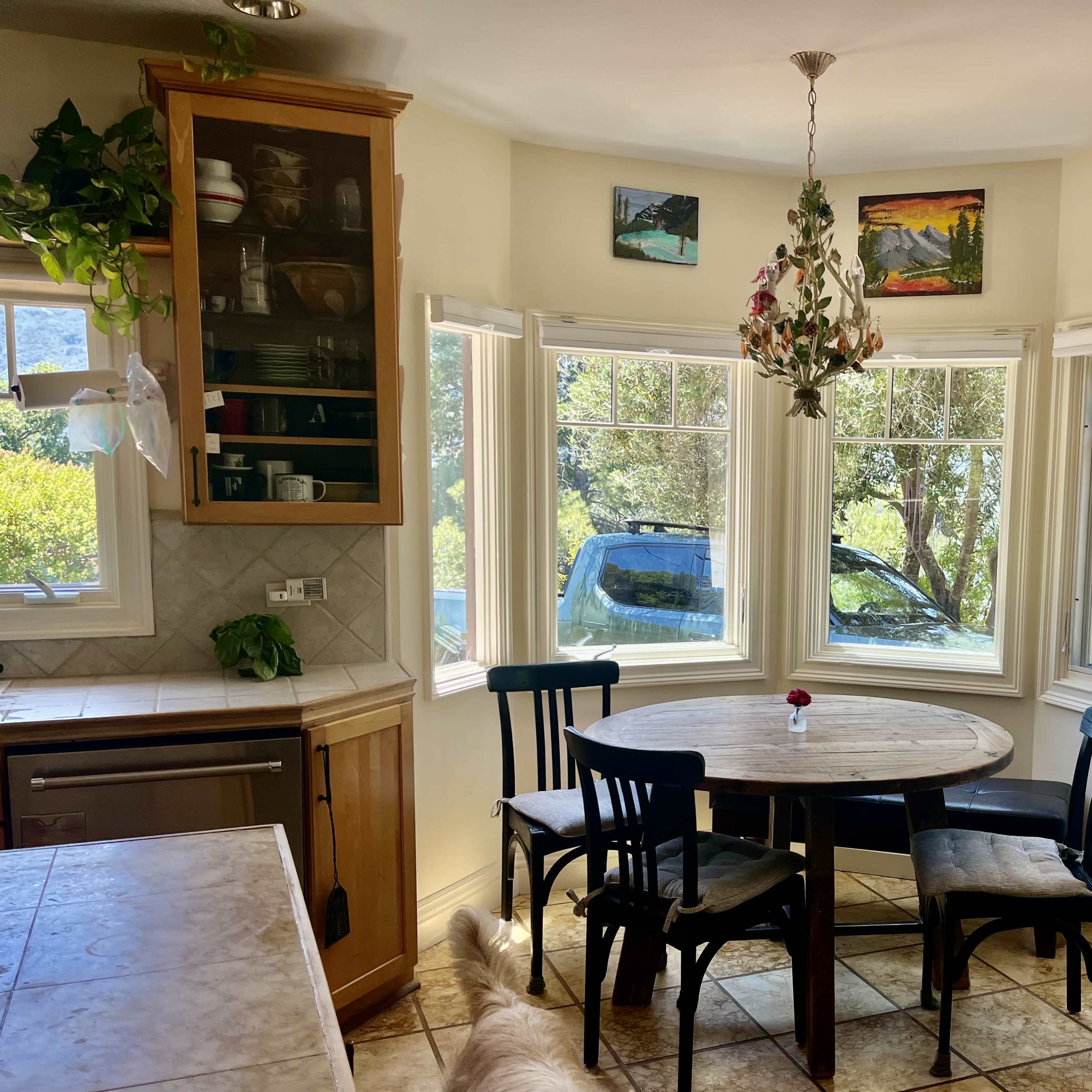 A kitchen features a round wooden table surrounded by four black chairs, with large windows displaying a view of greenery outside.