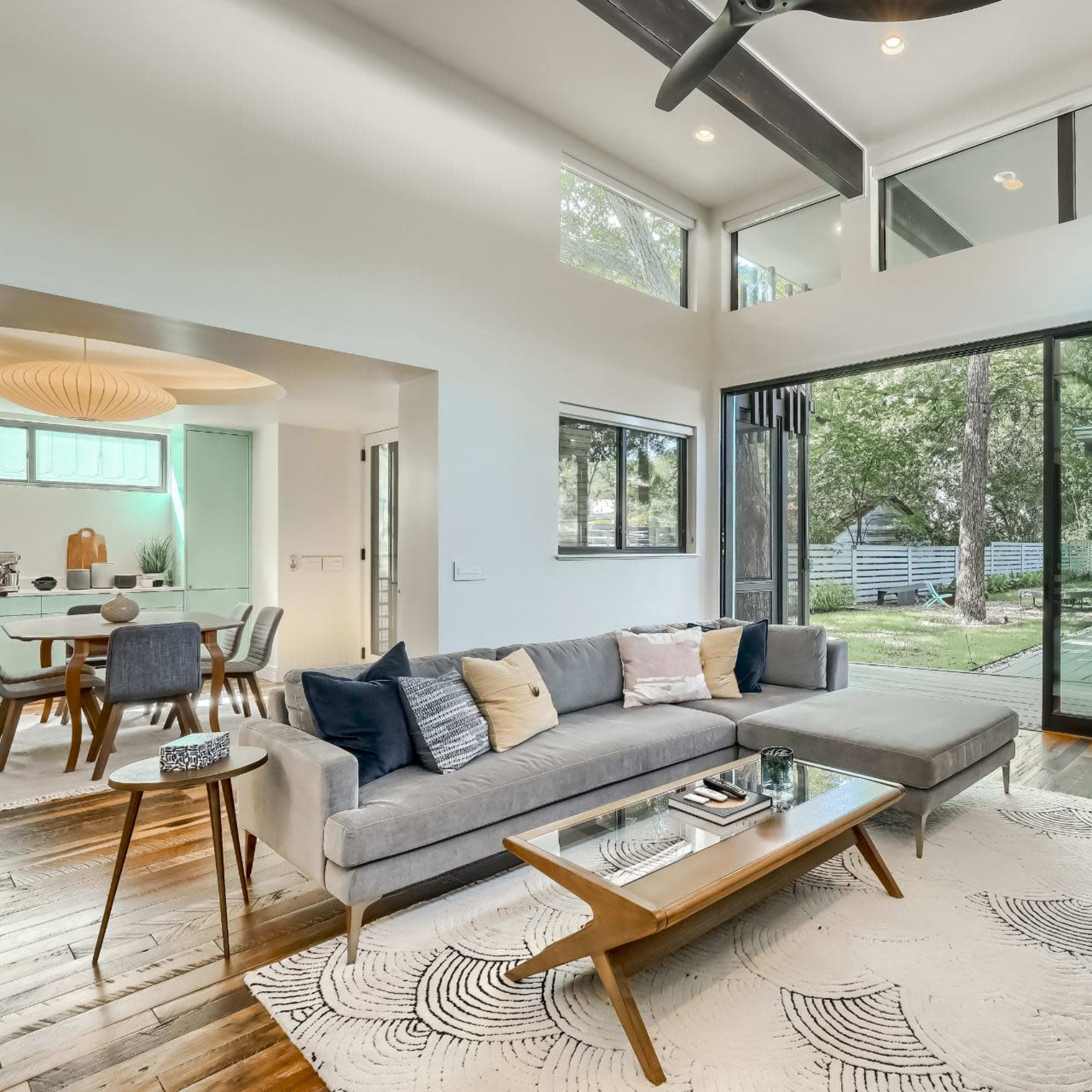 A modern living room featuring a gray sectional sofa, a wooden coffee table, and large windows overlooking a backyard with trees.