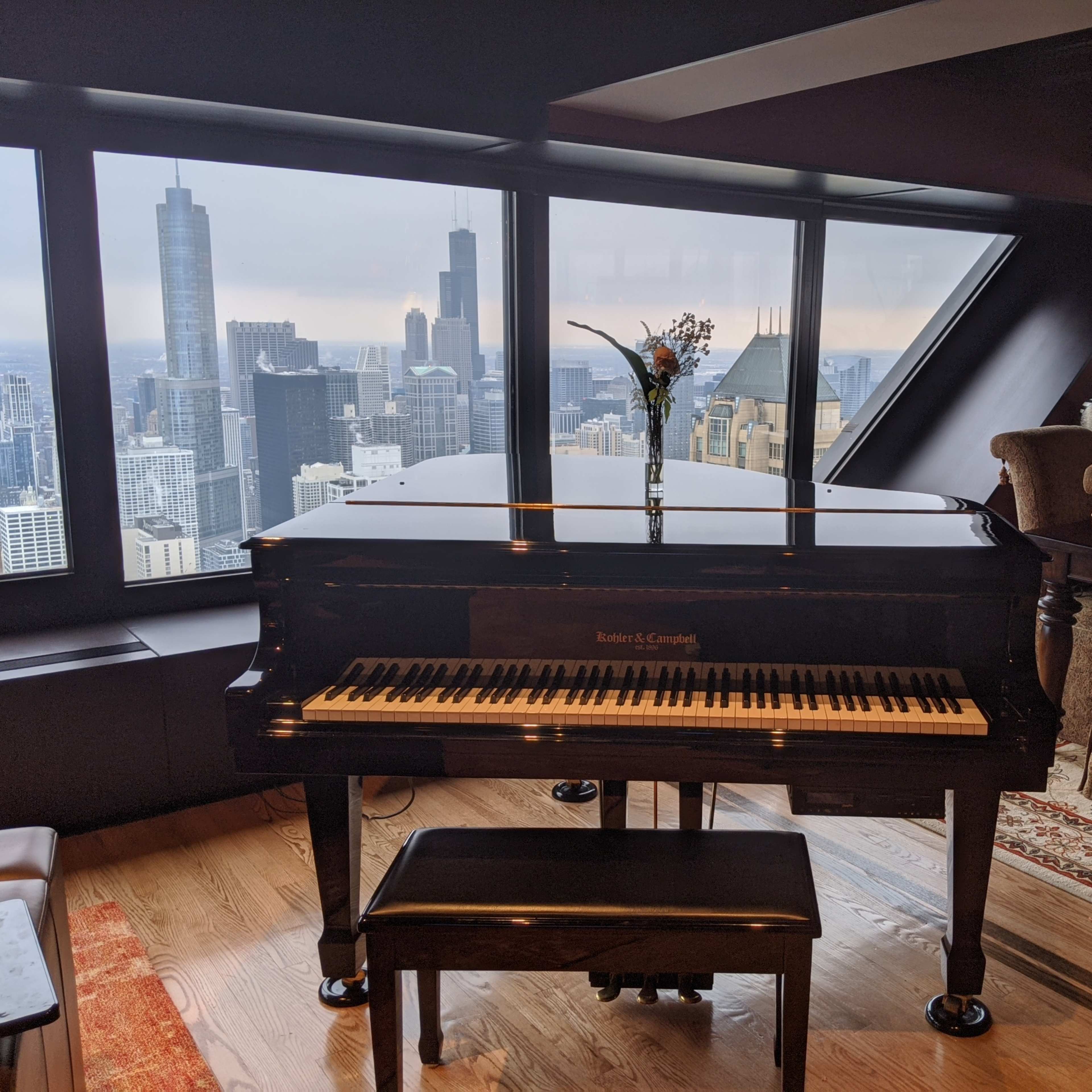 A grand piano with a vase of flowers sits in front of large windows overlooking a cityscape of skyscrapers.