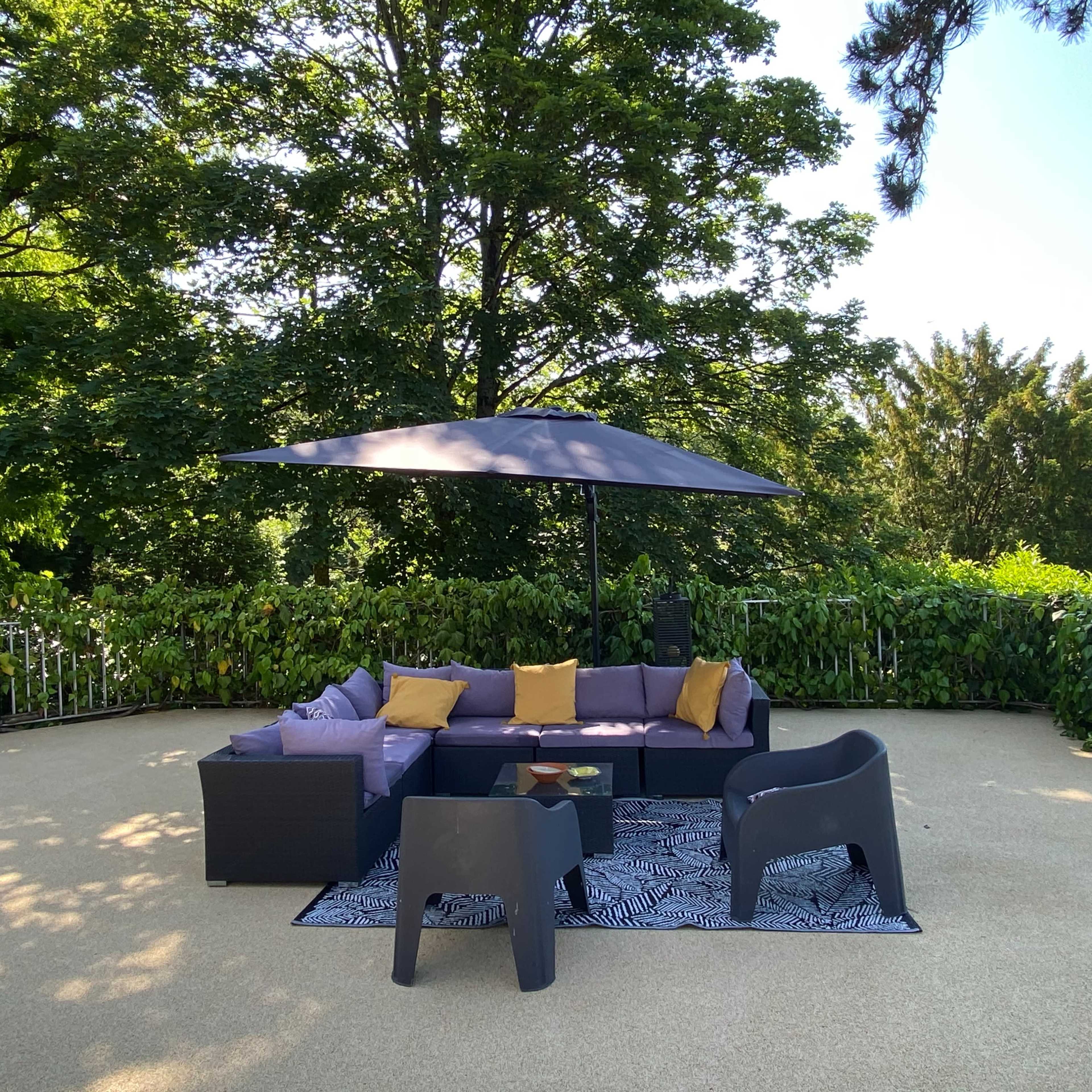 A patio area is furnished with a sectional sofa, two chairs, a coffee table, and a large umbrella, surrounded by greenery.