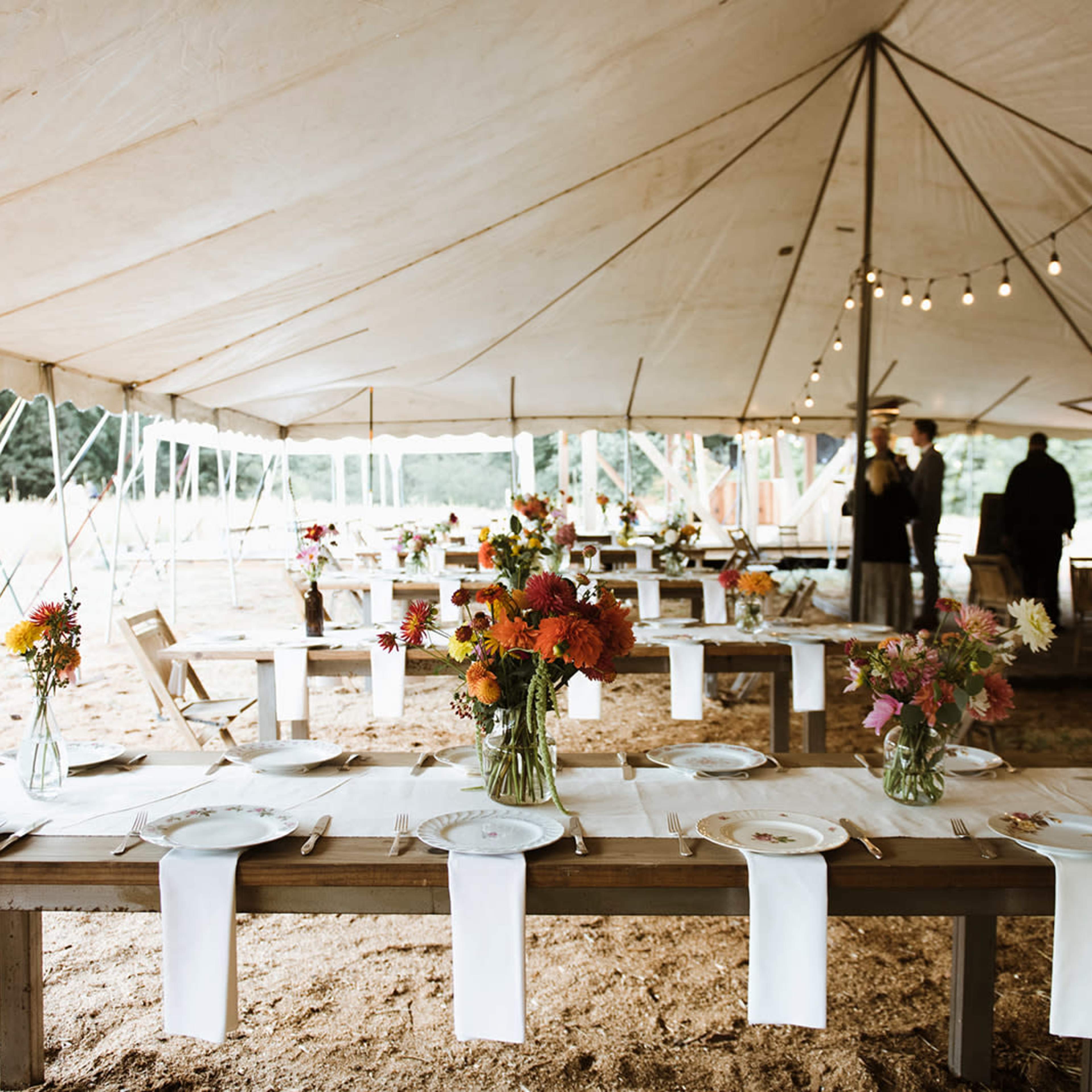 A spacious tent setup with wooden tables adorned with floral arrangements and simple white table runners is ready for an event.