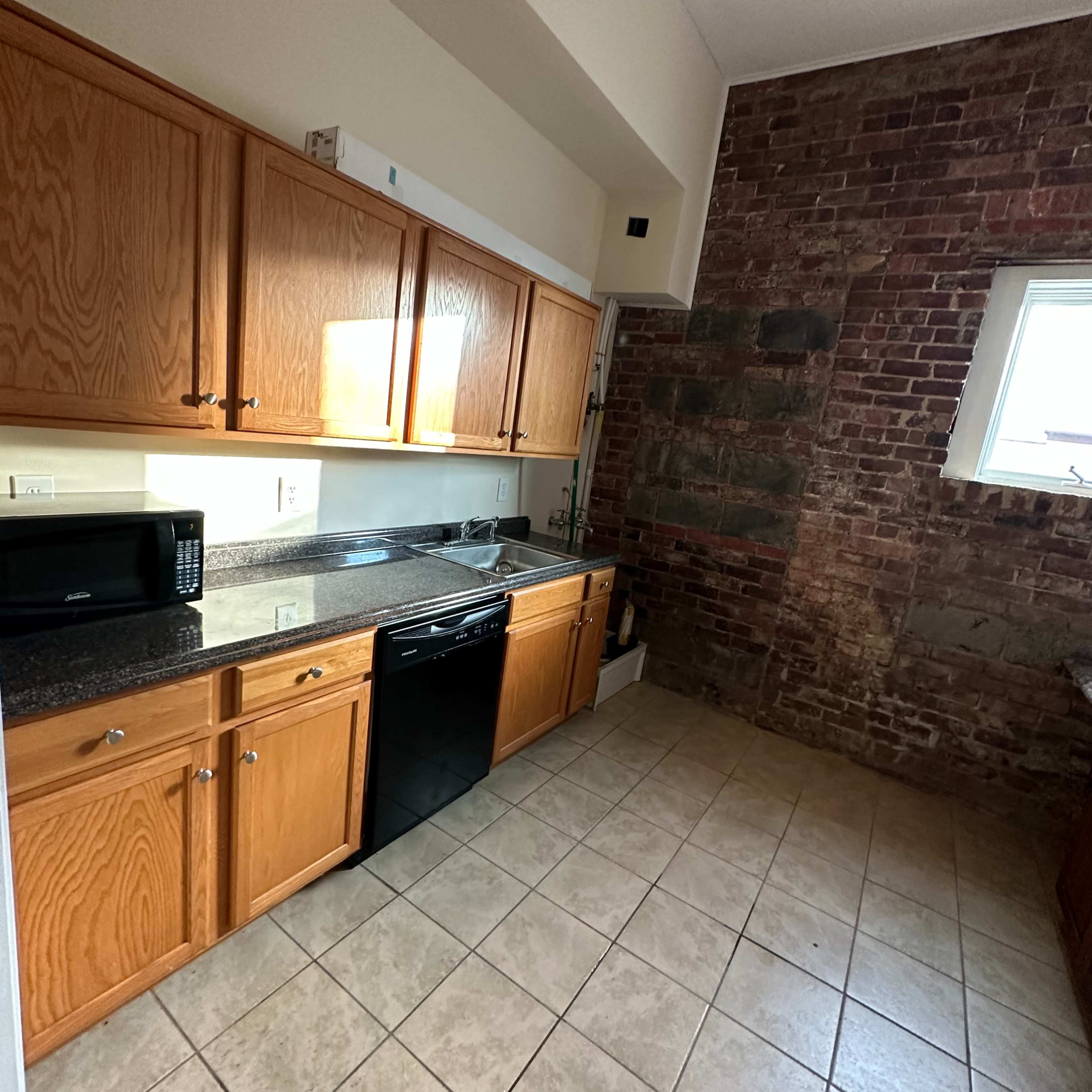 A kitchen with wooden cabinets, a black dishwasher, a microwave, and a brick wall.