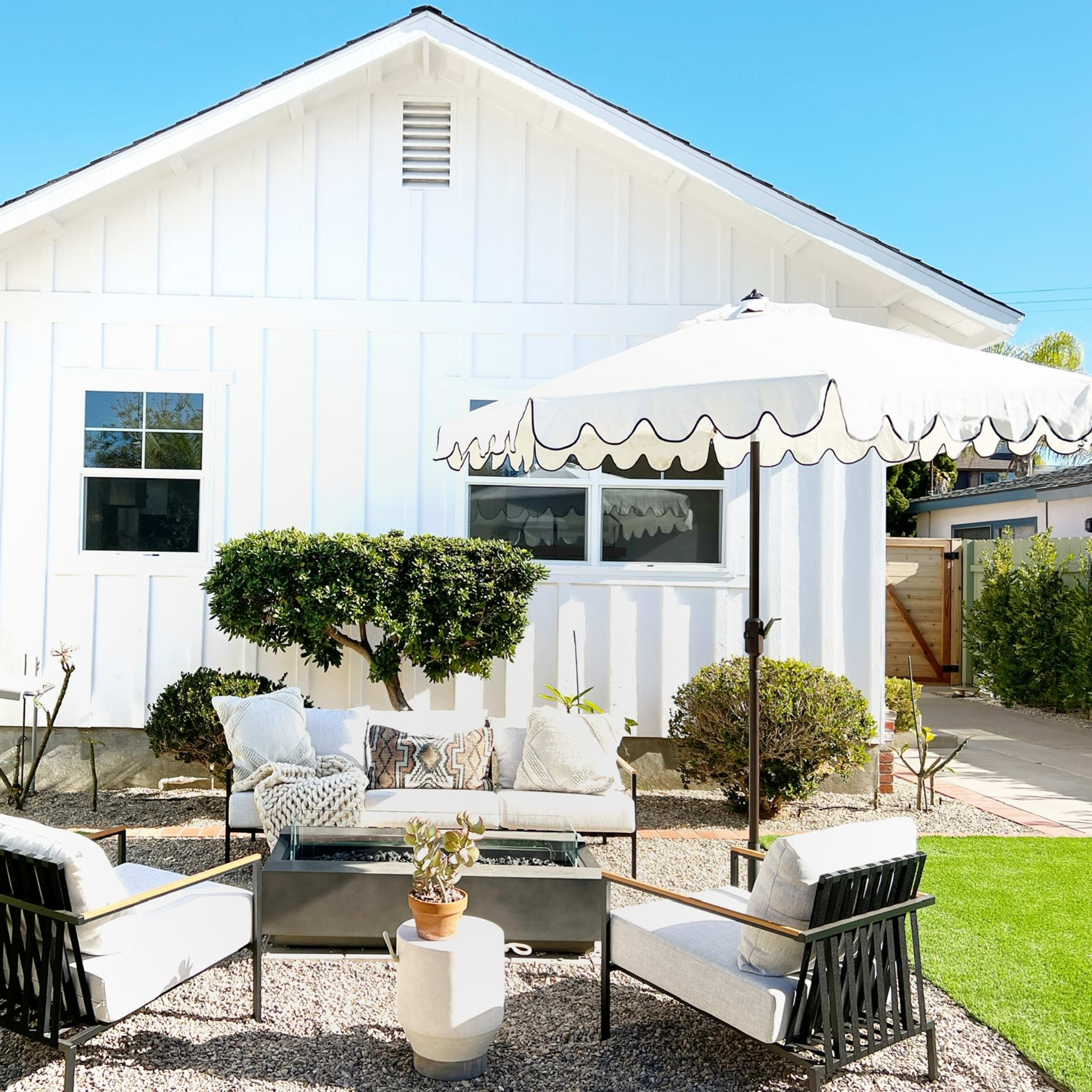 A backyard patio setup with a grill, seating area, and an umbrella near a white house.