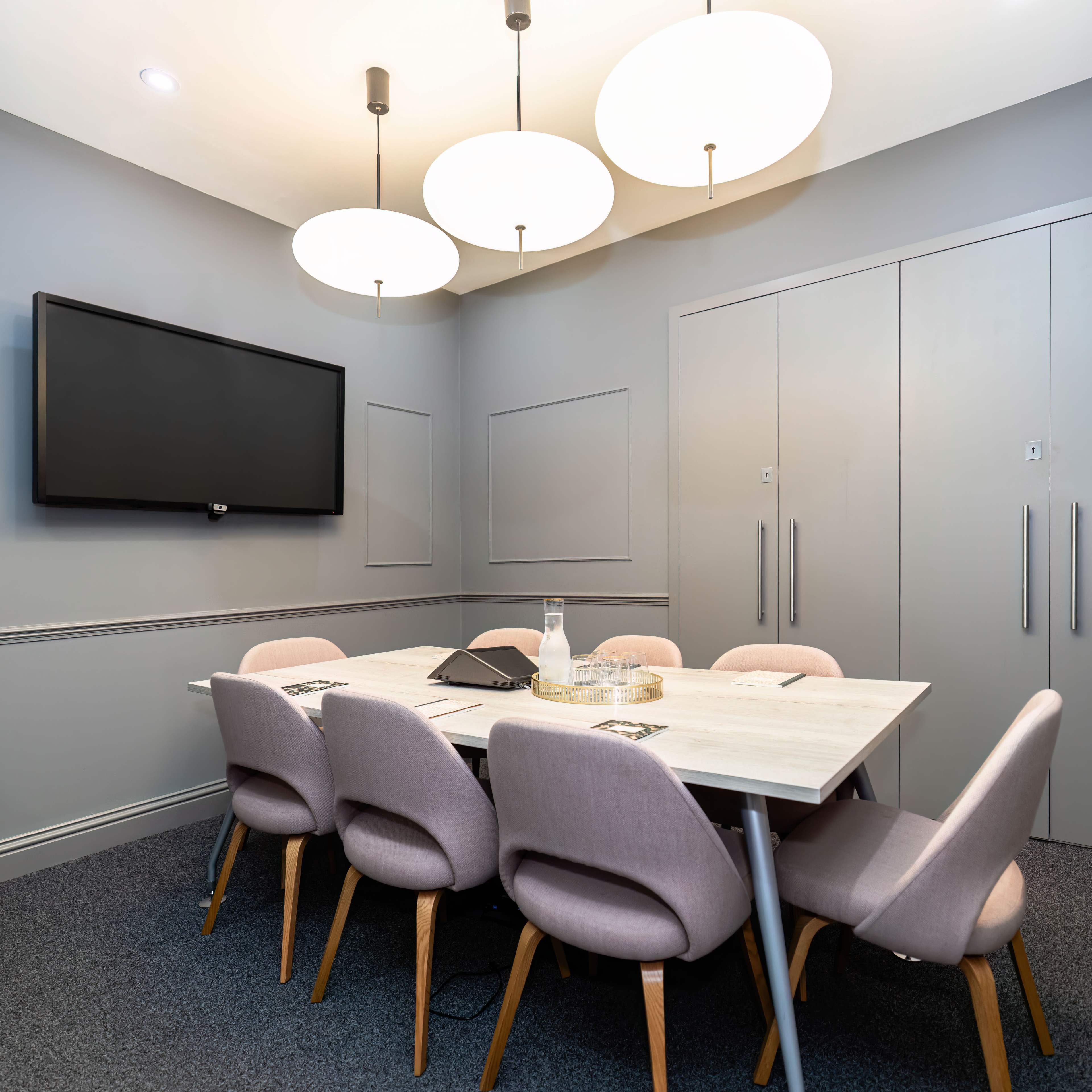 A modern meeting room features a table surrounded by six upholstered chairs, a wall-mounted TV, and large pendant lights overhead.