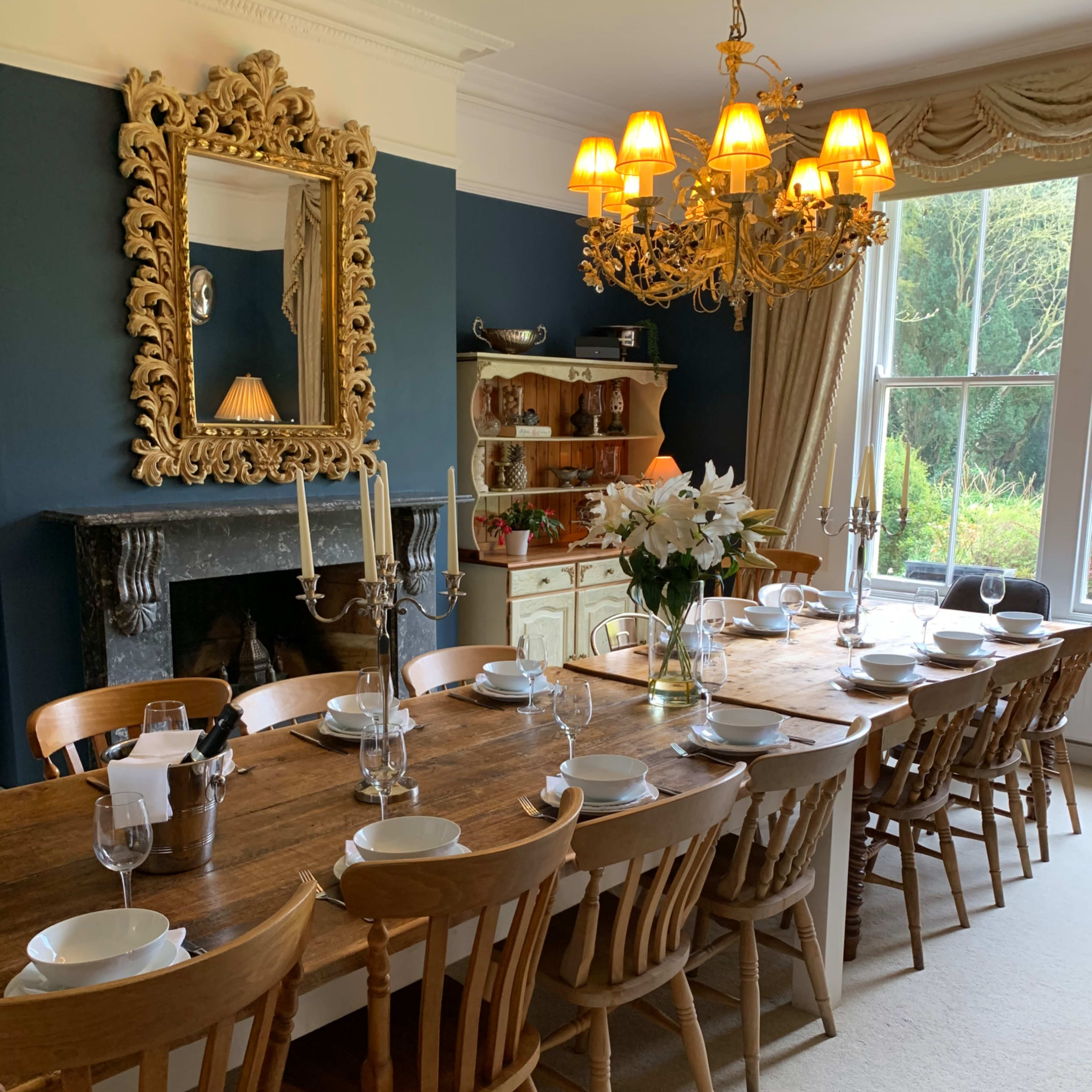 A long wooden dining table is set with plates and glasses, surrounded by wooden chairs in a room featuring a large ornate mirror and a chandelier.