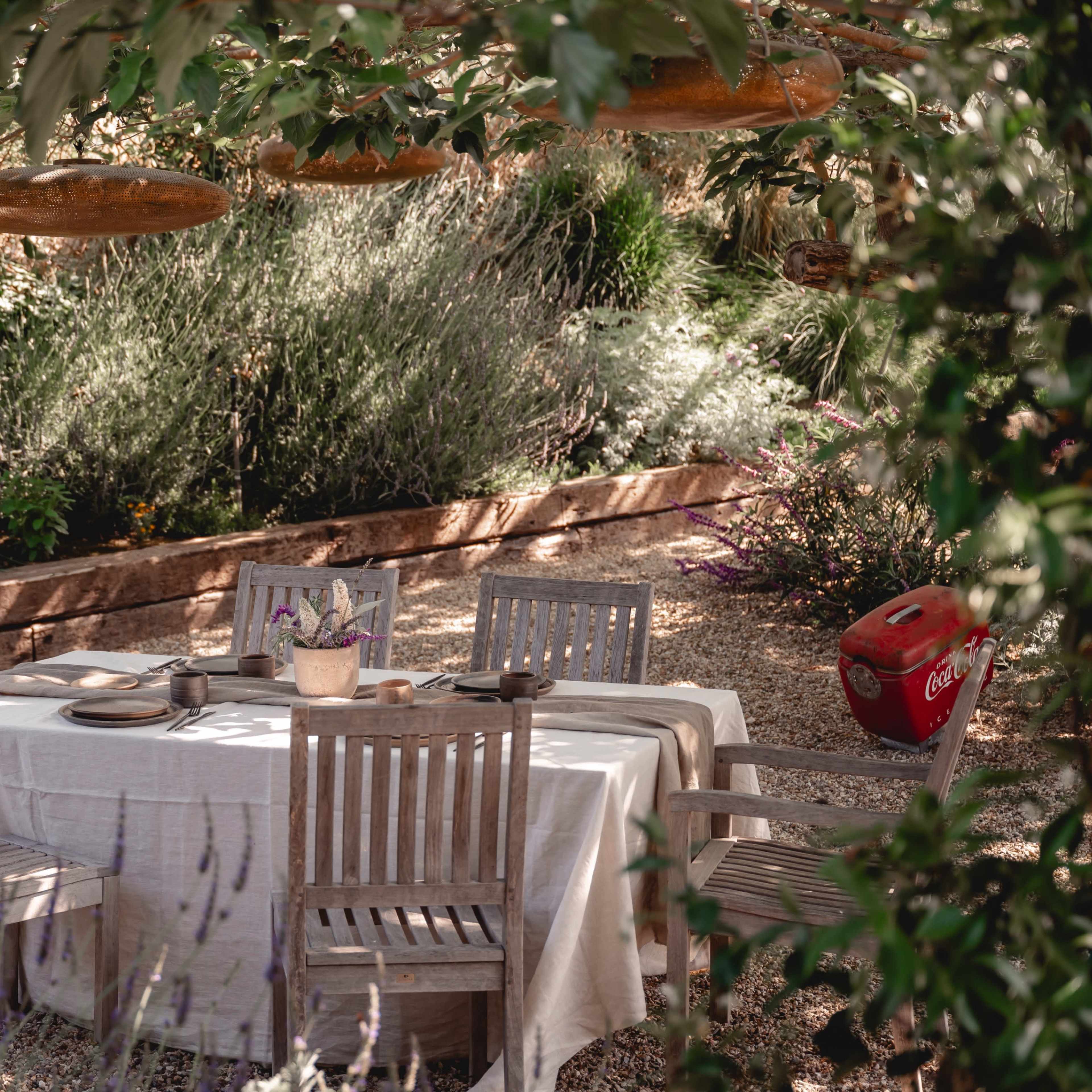 A wooden table is set with plates and utensils, surrounded by rustic chairs under greenery in a garden.