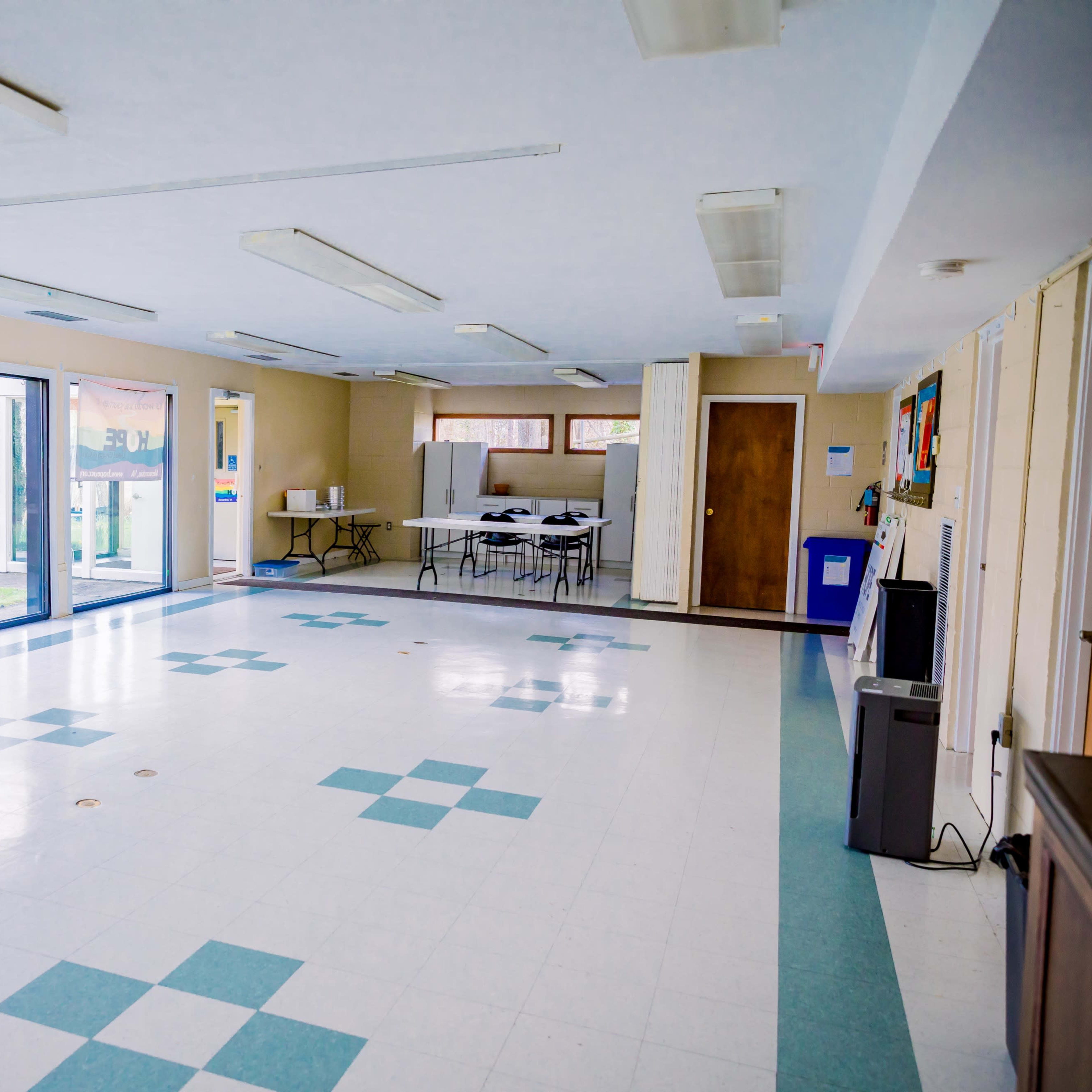 A spacious, empty room with tiled flooring, large windows, and tables set up against the wall.