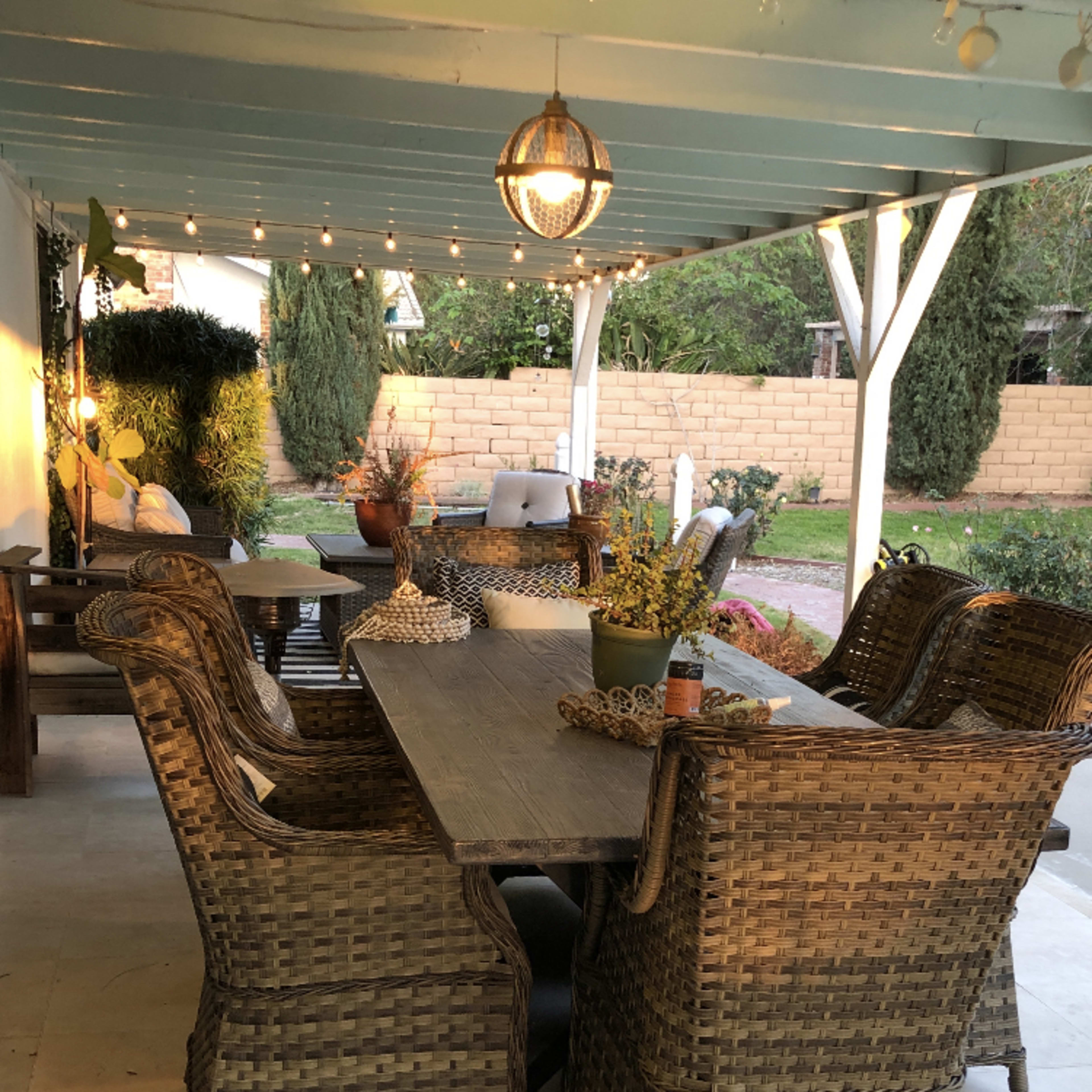 A covered outdoor dining area features a long table surrounded by wicker chairs, with string lights hanging above and a garden visible in the background.