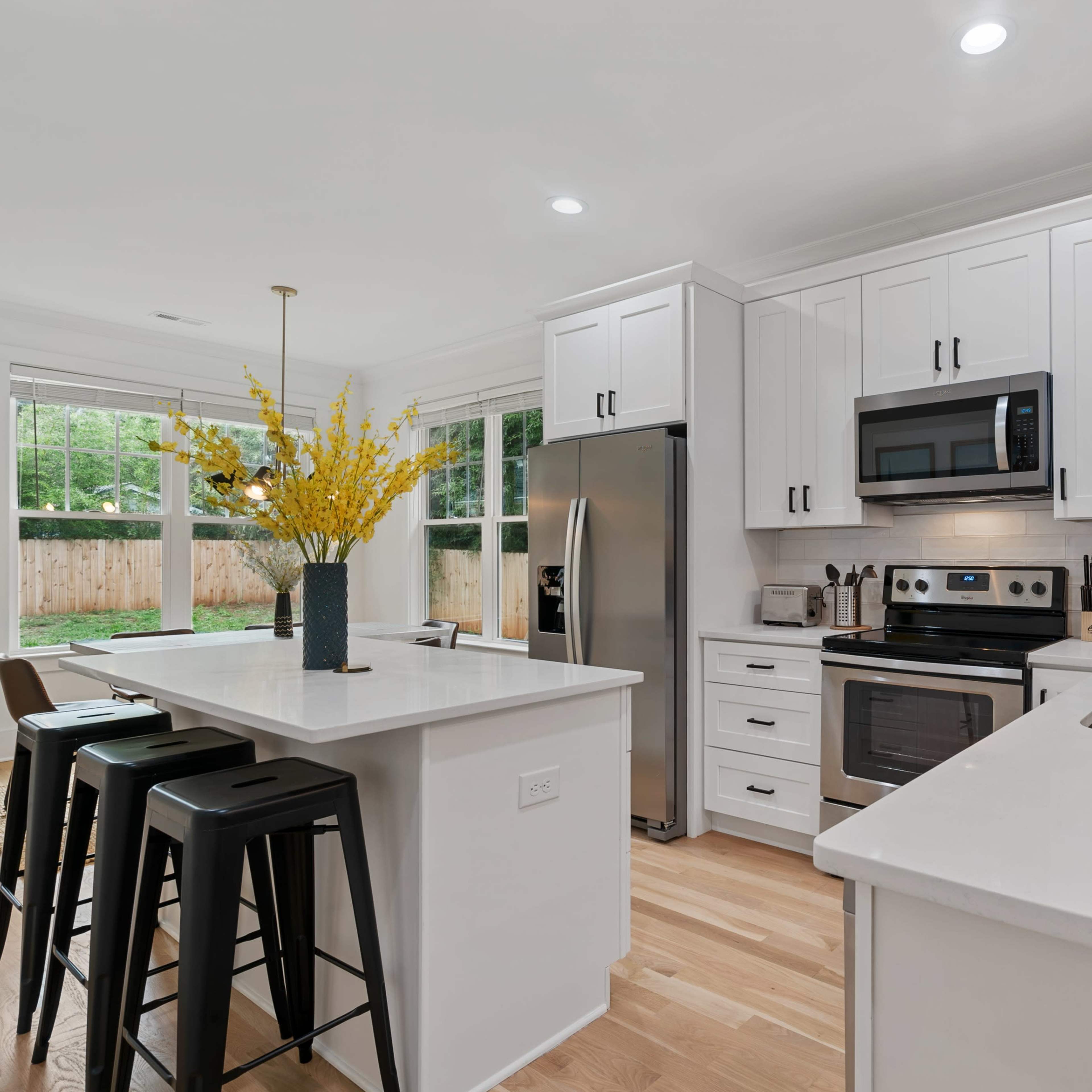 A modern kitchen with white cabinetry, stainless steel appliances, and a central island featuring black stools and a vase of yellow flowers.