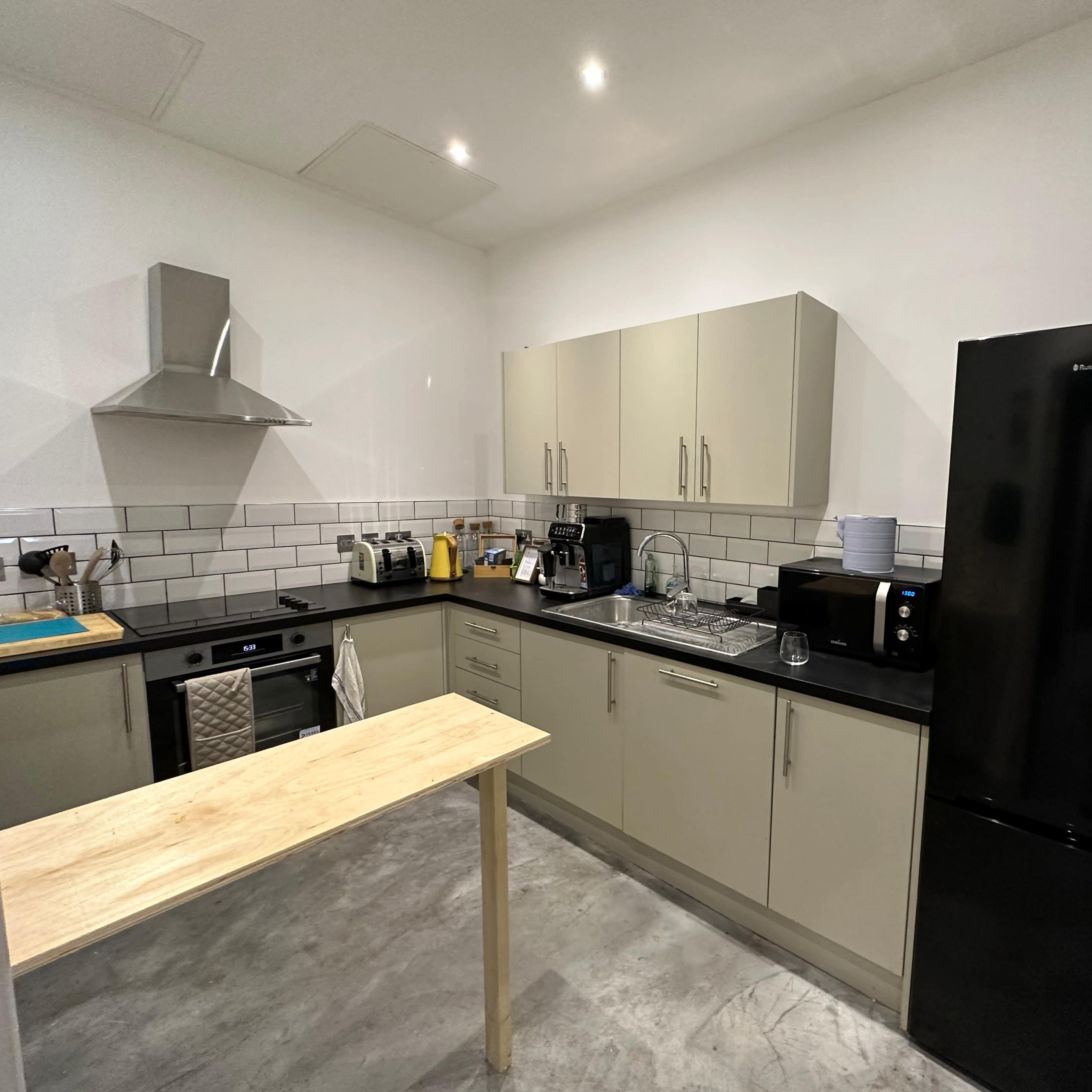 A modern kitchen with light cabinetry, a black refrigerator, a stainless steel oven, and a wooden table in the center.
