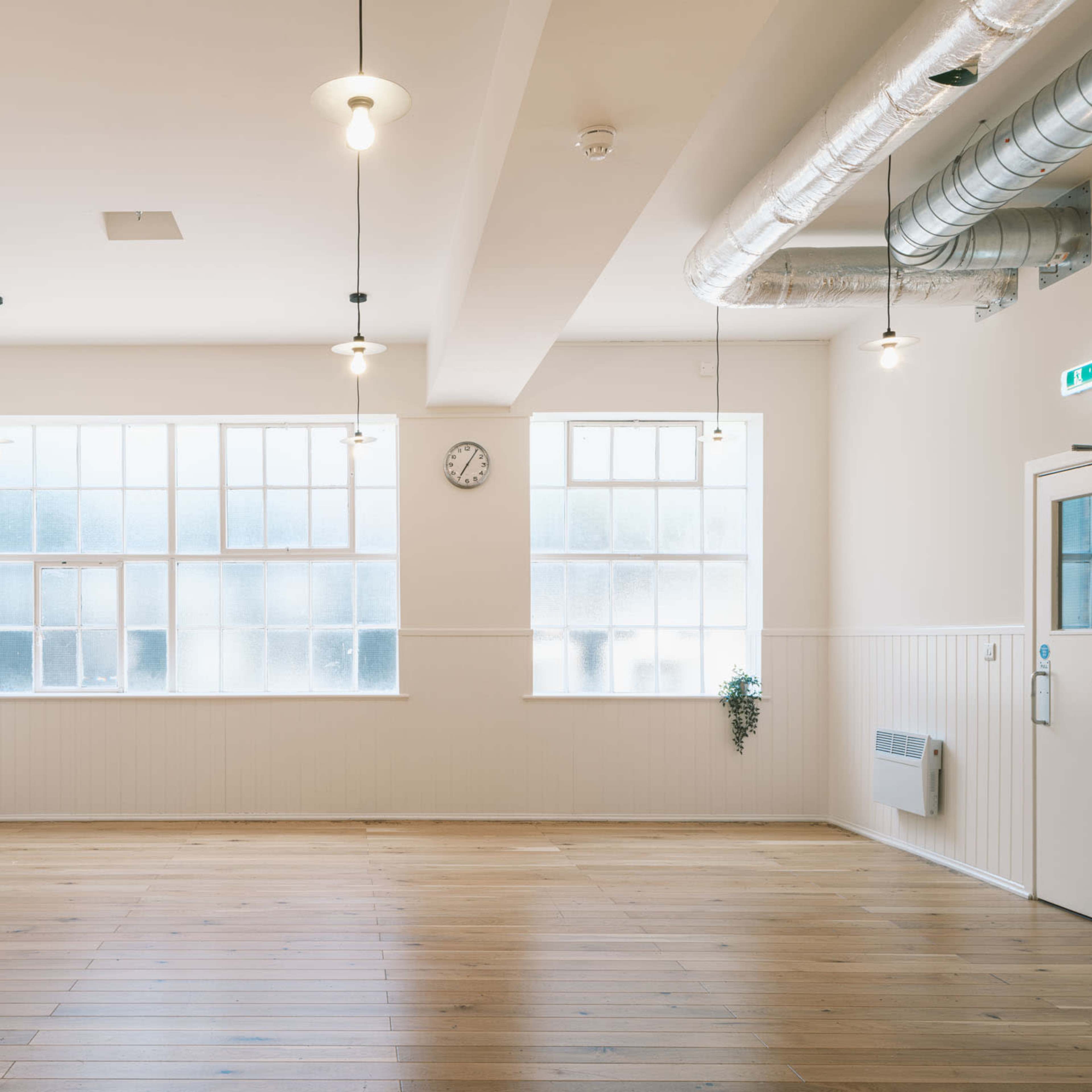 A spacious, empty room with wooden flooring, large windows, and industrial-style lighting fixtures.