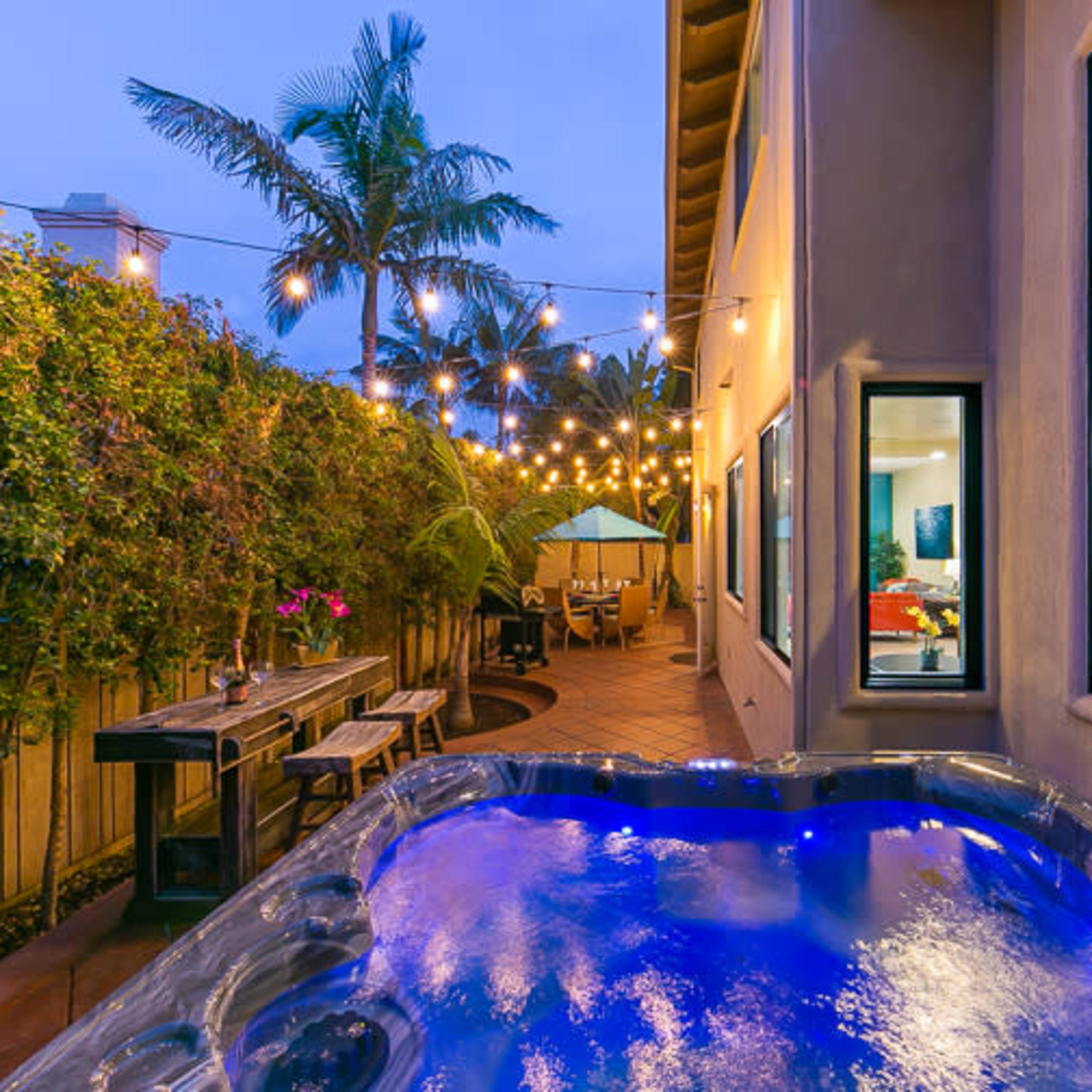 A backyard with a hot tub illuminated by blue lights, string lights overhead, and a dining area in the background surrounded by greenery.