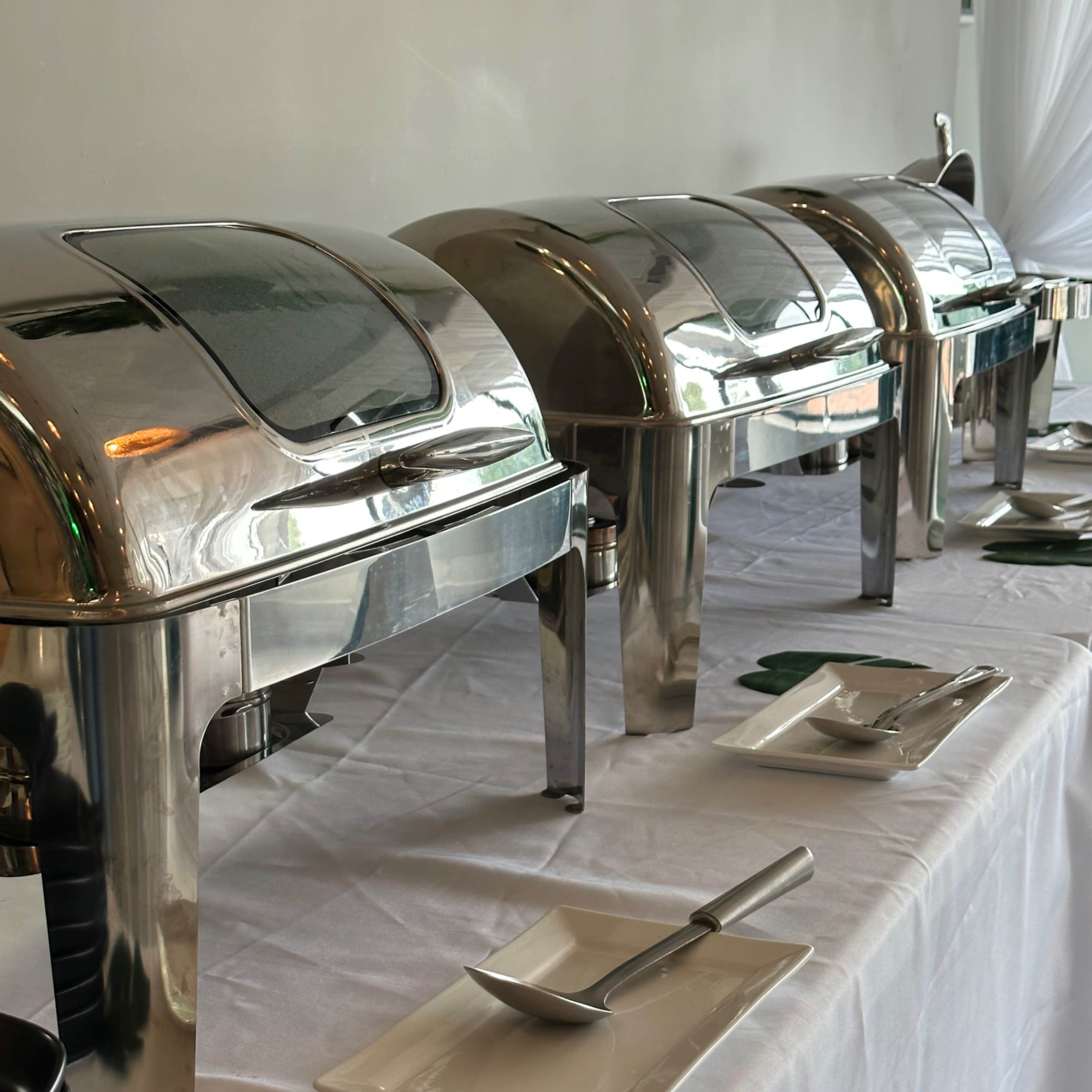 A row of stainless steel chafing dishes on a table covered with a white tablecloth, accompanied by white plates and utensils.