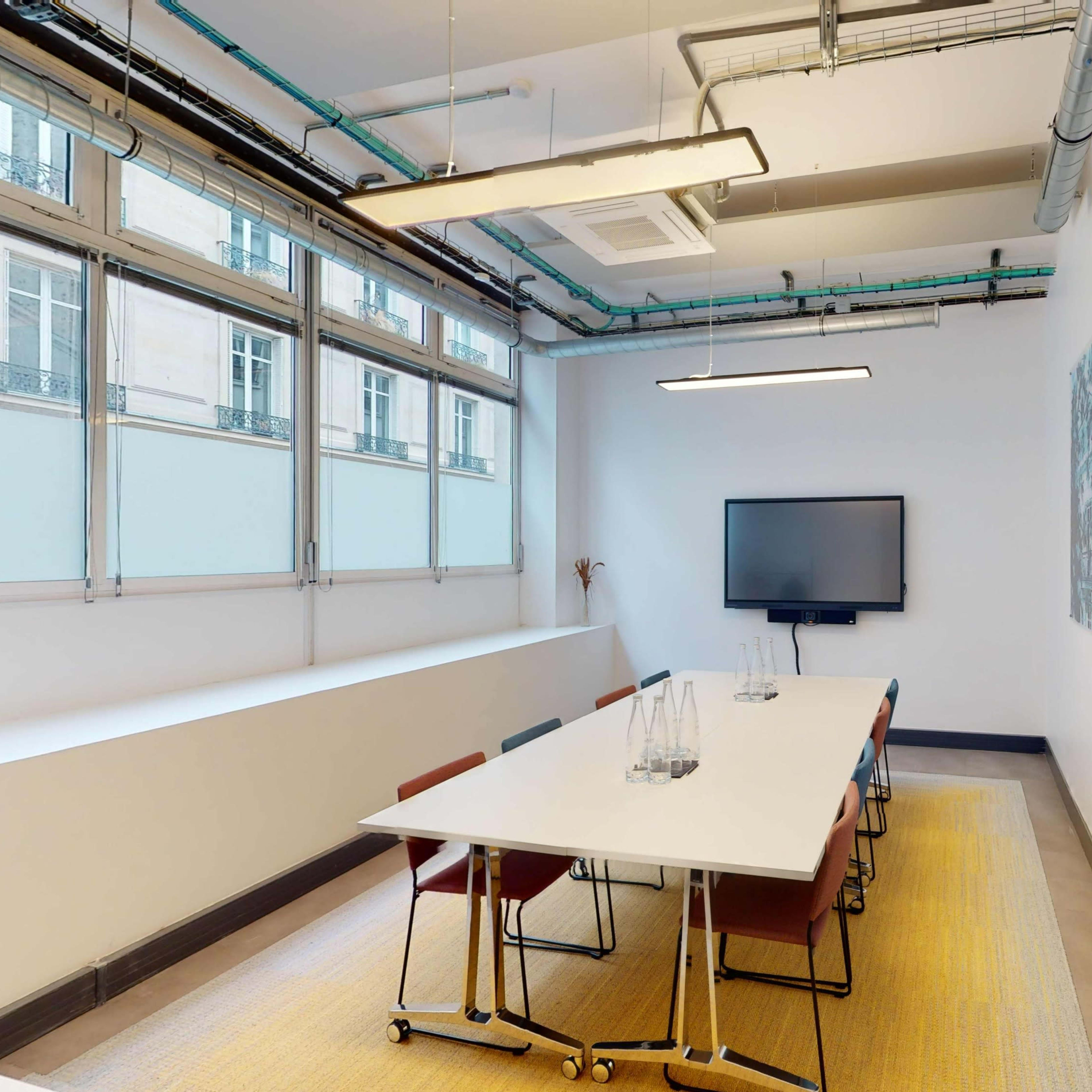 A meeting room features a long white table surrounded by chairs, a TV on the wall, and large windows allowing natural light.