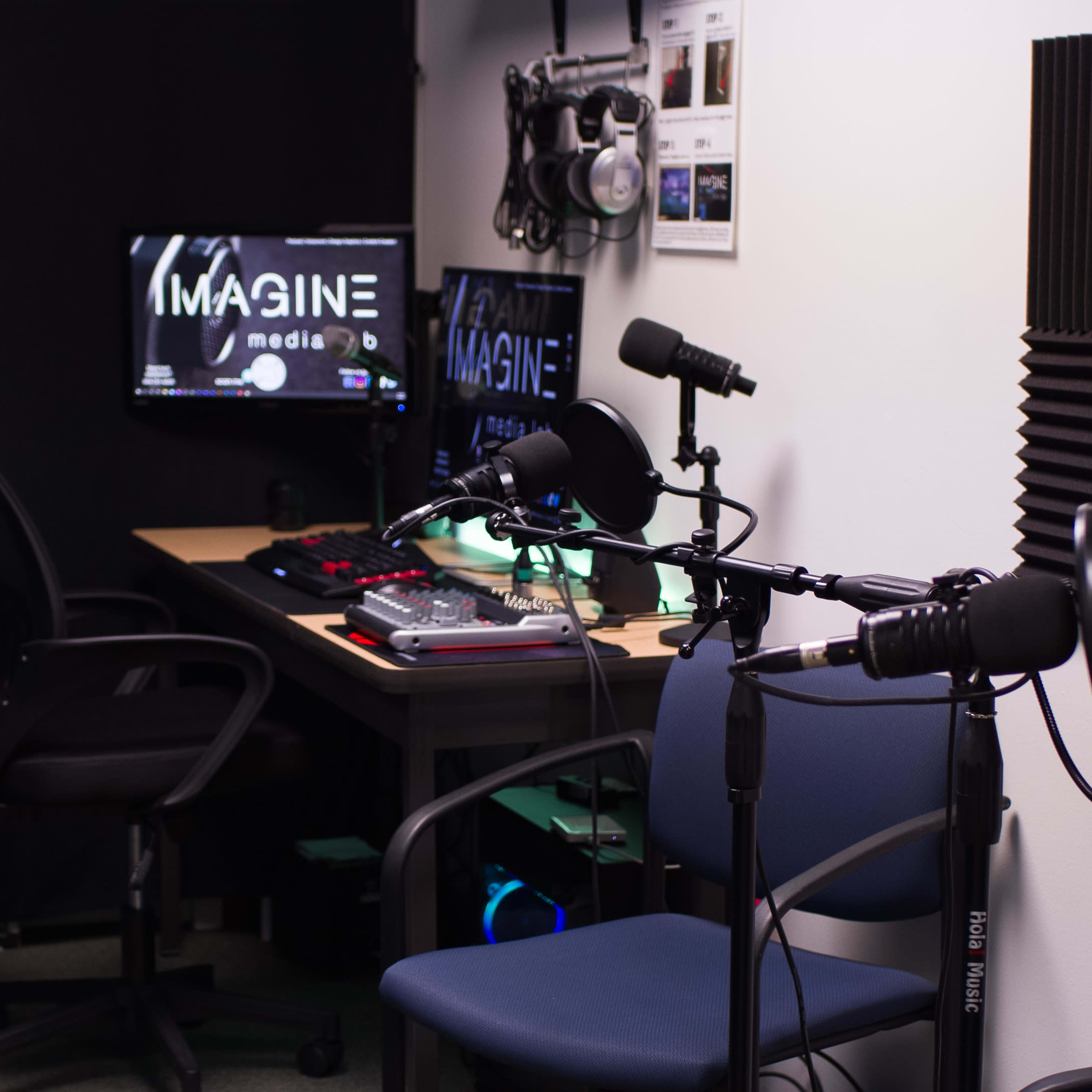A small recording studio with a desk featuring a computer and audio equipment, chairs, and a black soundproof wall.
