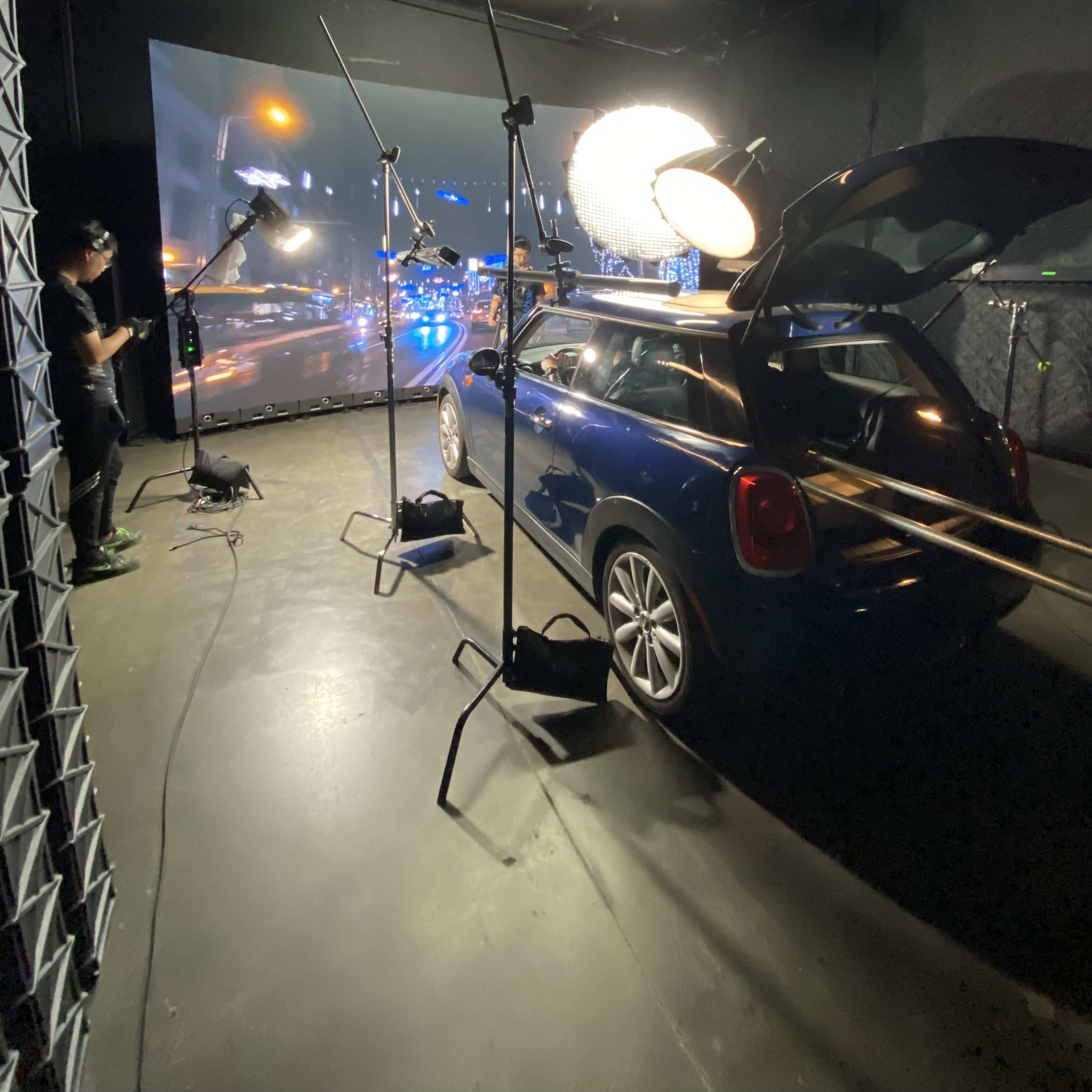 A blue car is positioned inside a studio with lighting equipment, while a large screen displays a night cityscape in the background.