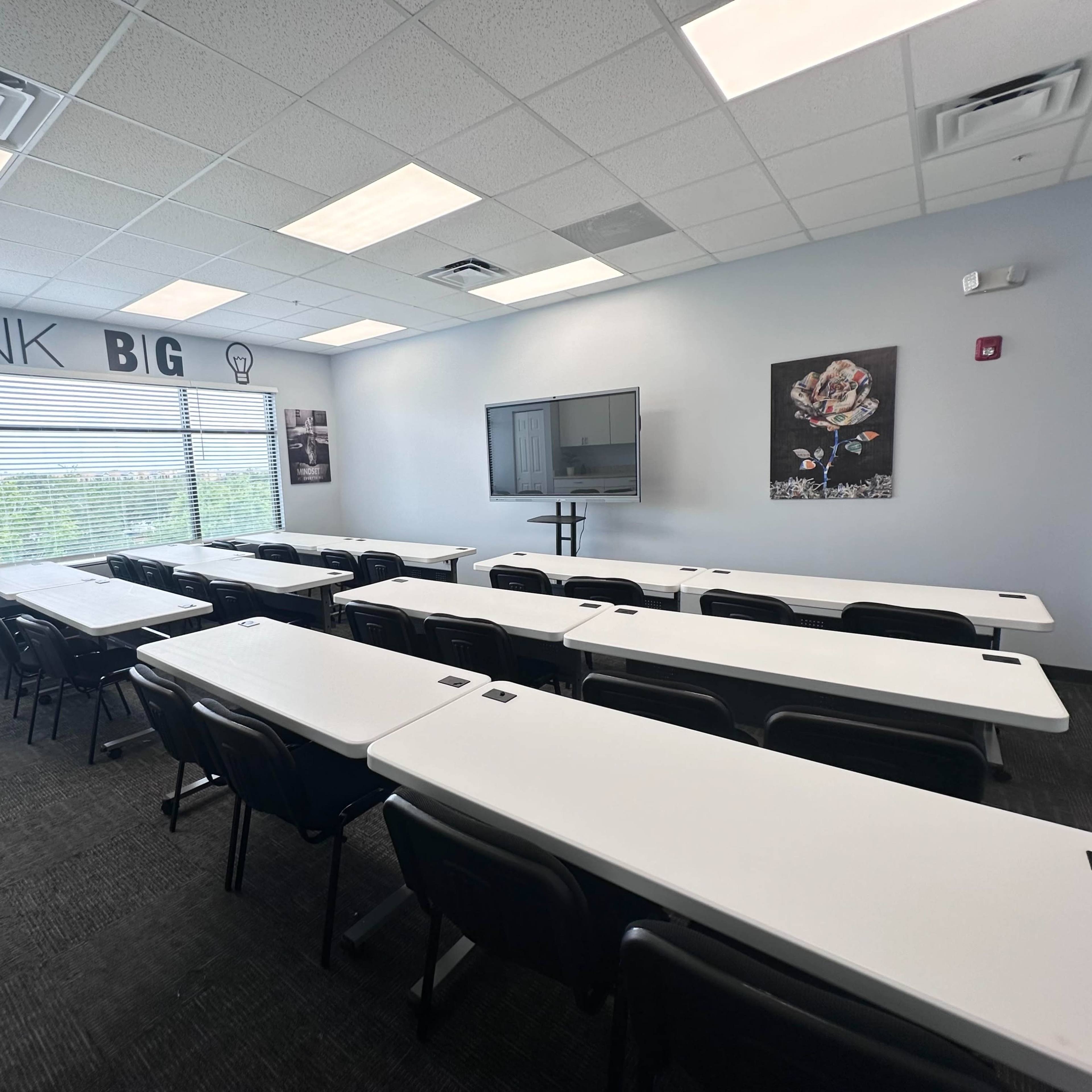 A modern classroom is set up with multiple tables and chairs arranged in rows, featuring large windows and a wall with motivational text.