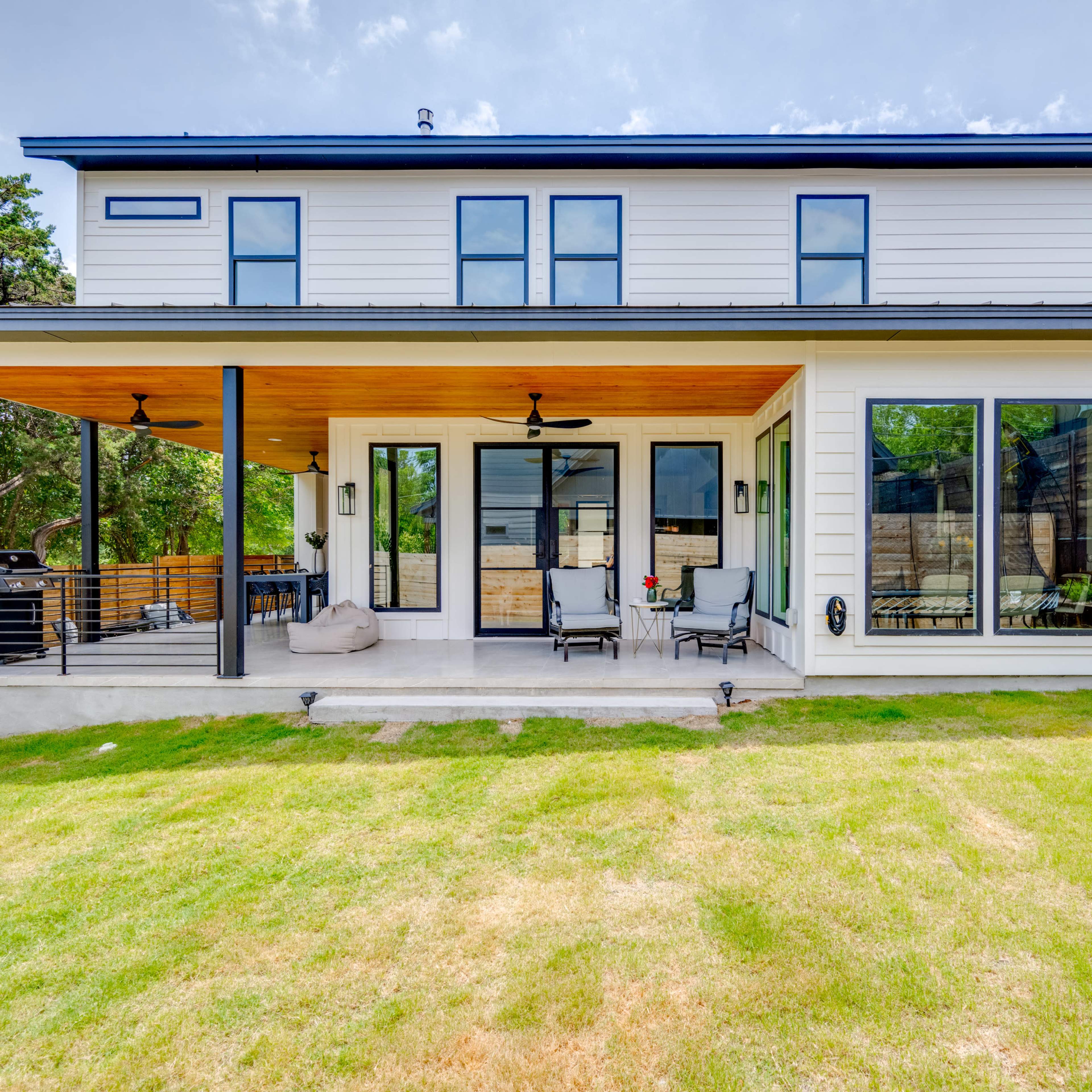 A modern two-story house with large windows and a wooden deck, surrounded by greenery.