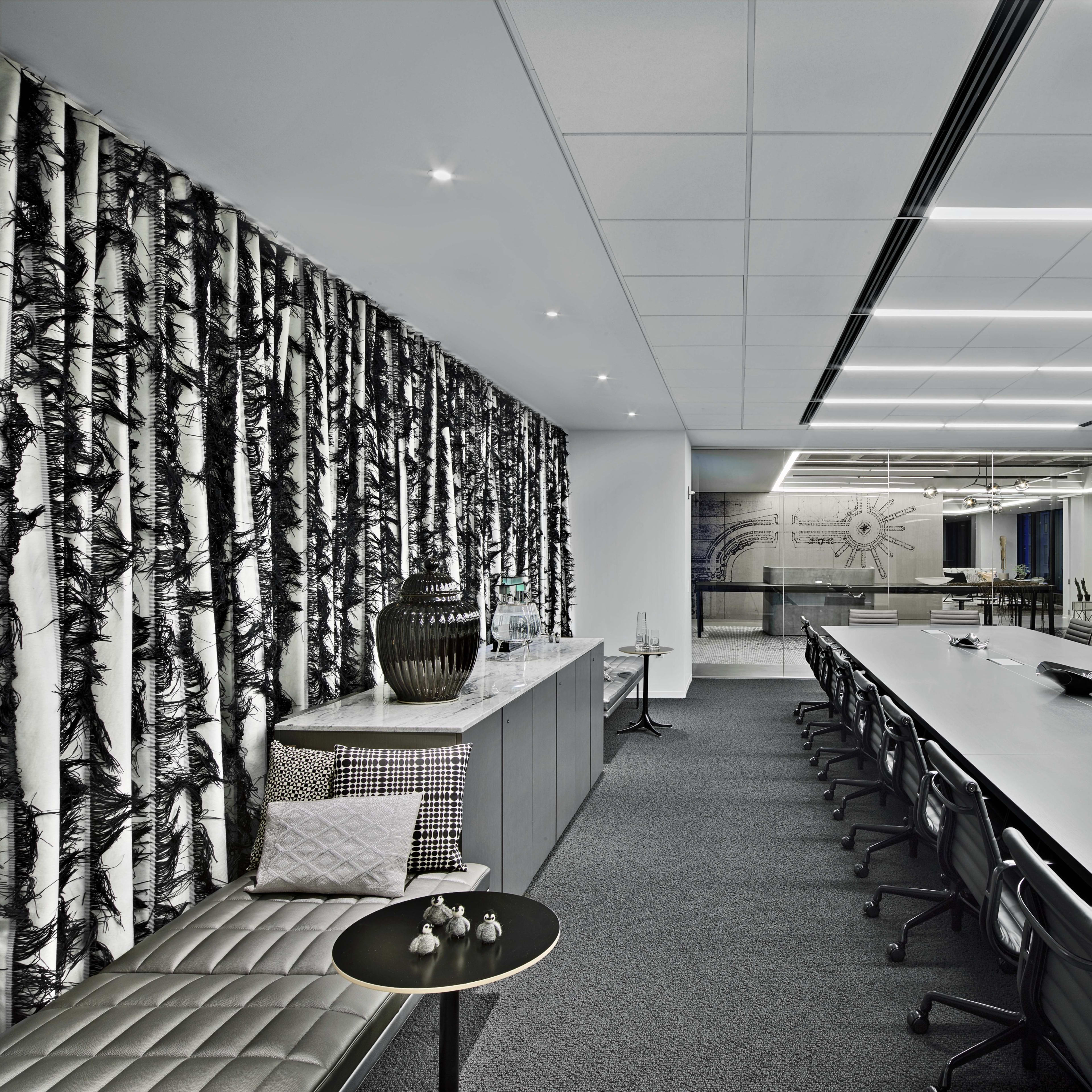 A modern conference room features a long table with black chairs, a textured wall adorned with a tree design, and a glass-walled area displaying additional seating.
