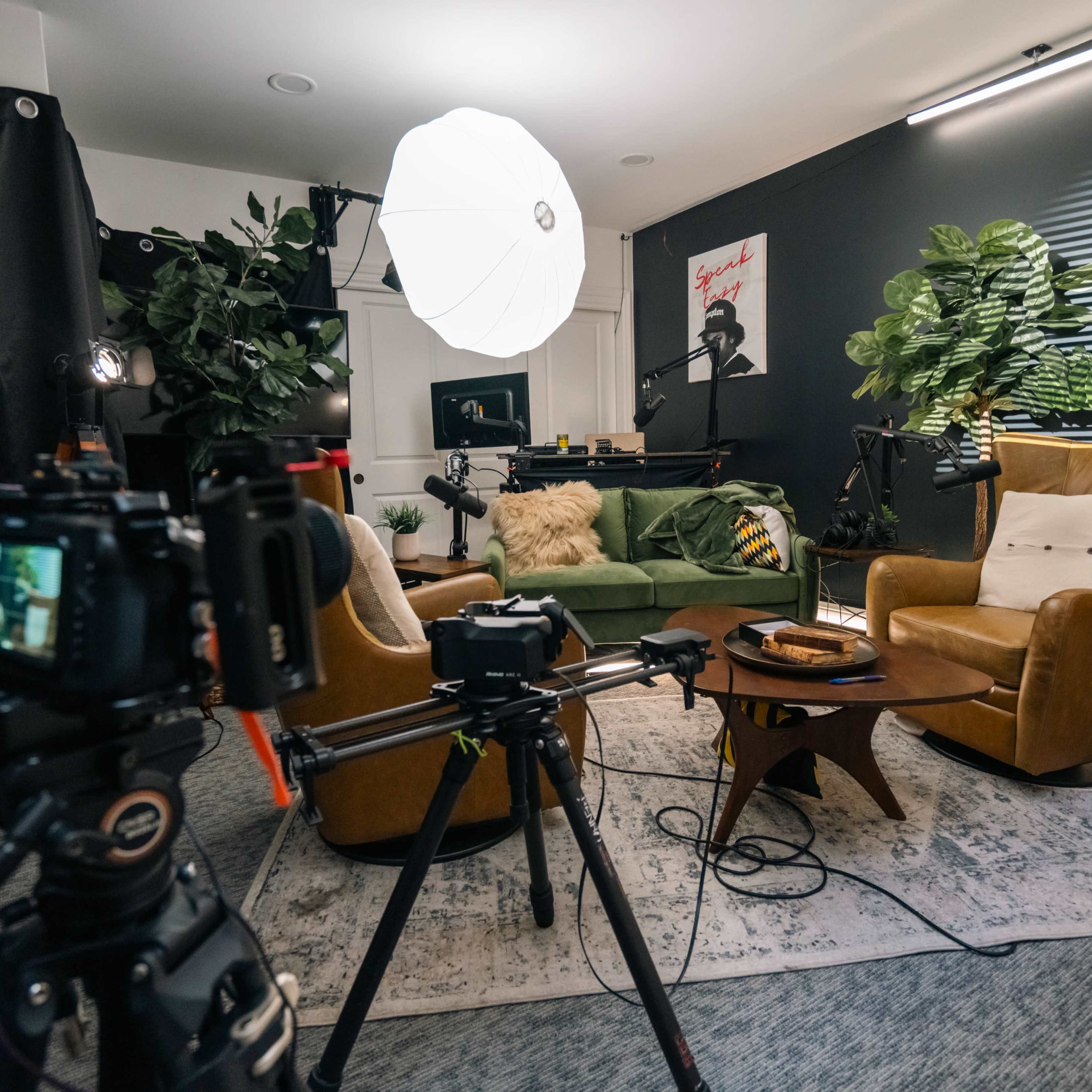 A well-arranged indoor studio setup with a green sofa, two armchairs, and various recording equipment, surrounded by plants and posters.