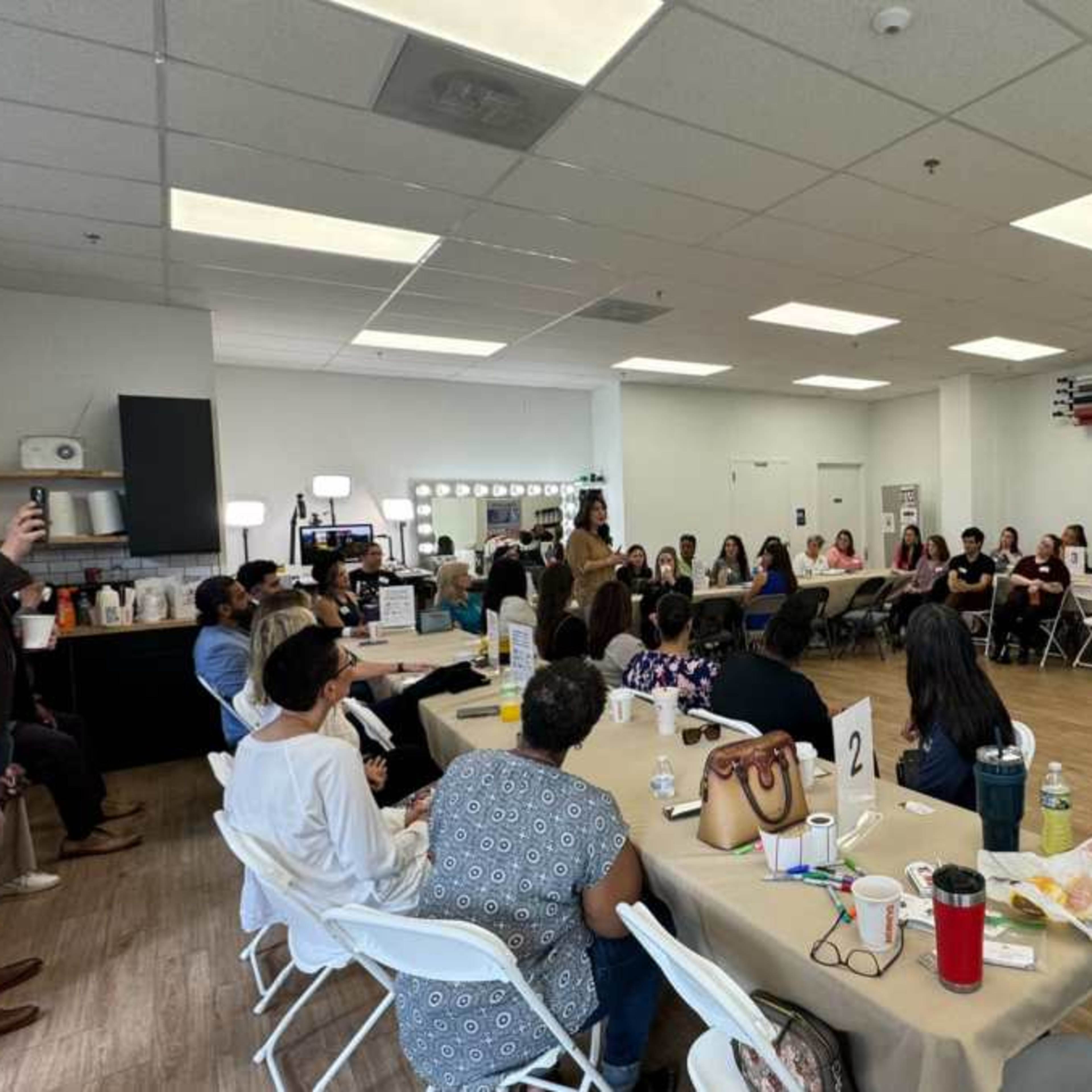 A large group of people is gathered in a well-lit room, sitting at tables and listening to a speaker at the front.