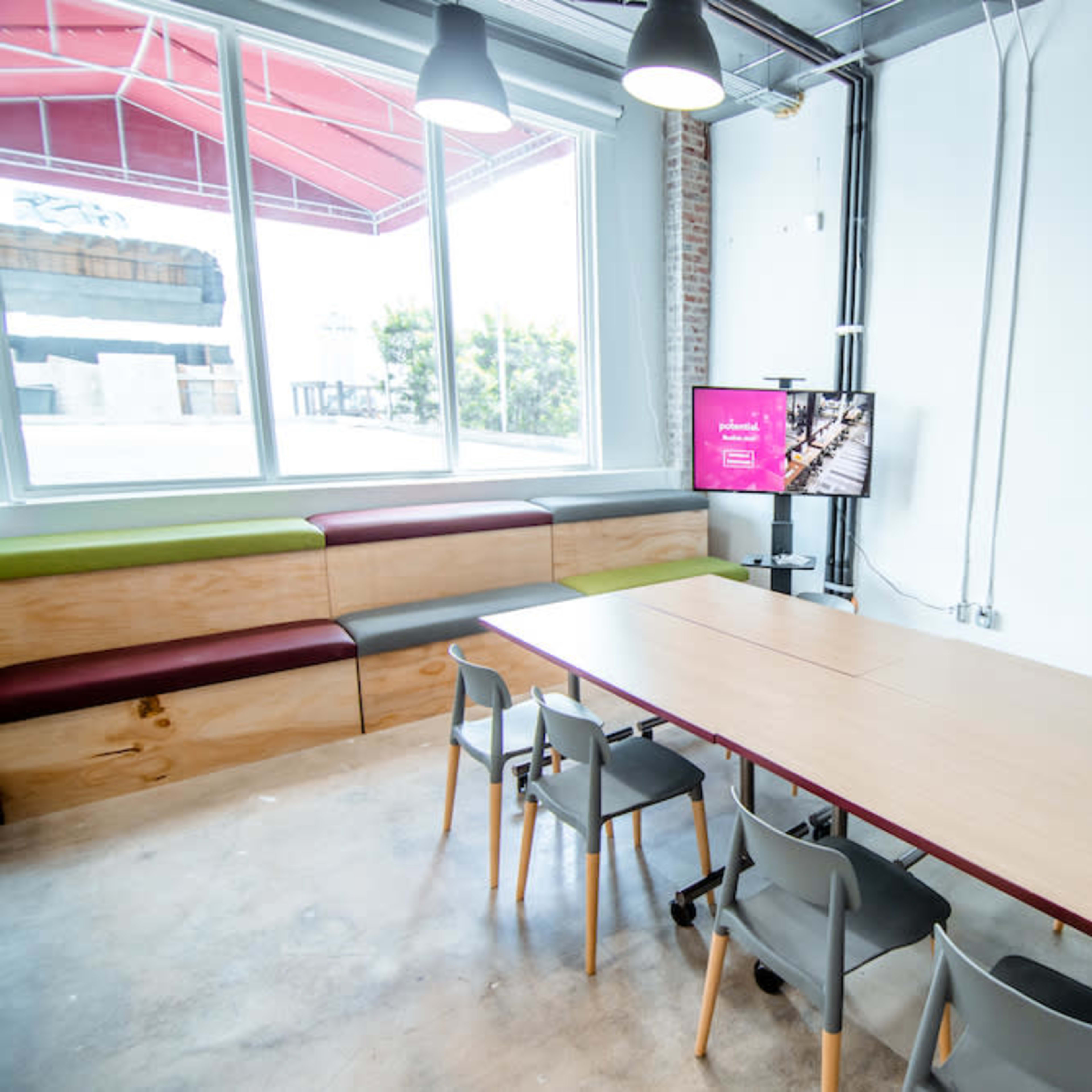 A modern conference room features a long wooden table surrounded by chairs, with a large window and a television display on one wall.