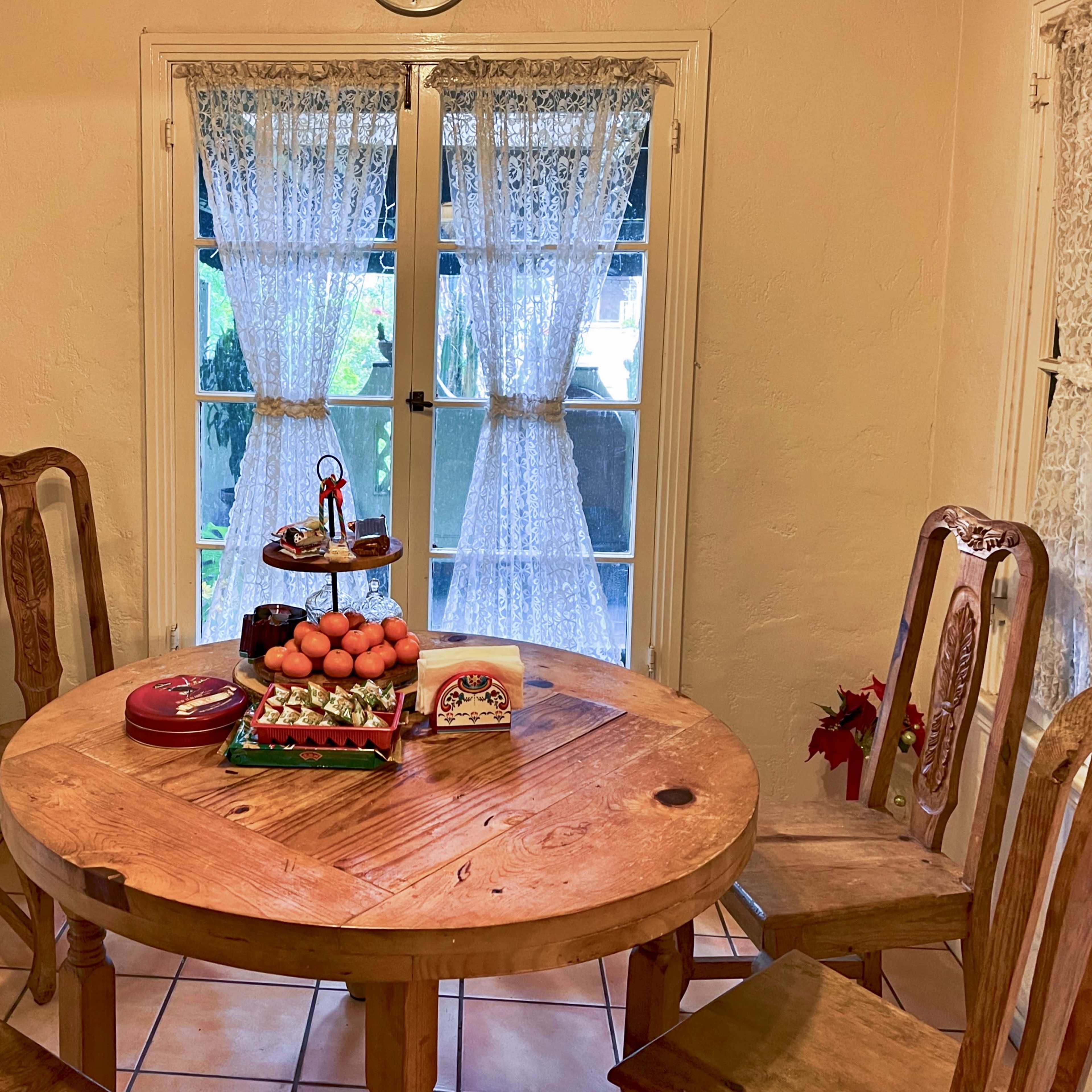 A wooden dining table with a plate of fruit and desserts is set in a well-lit room near lace curtains.