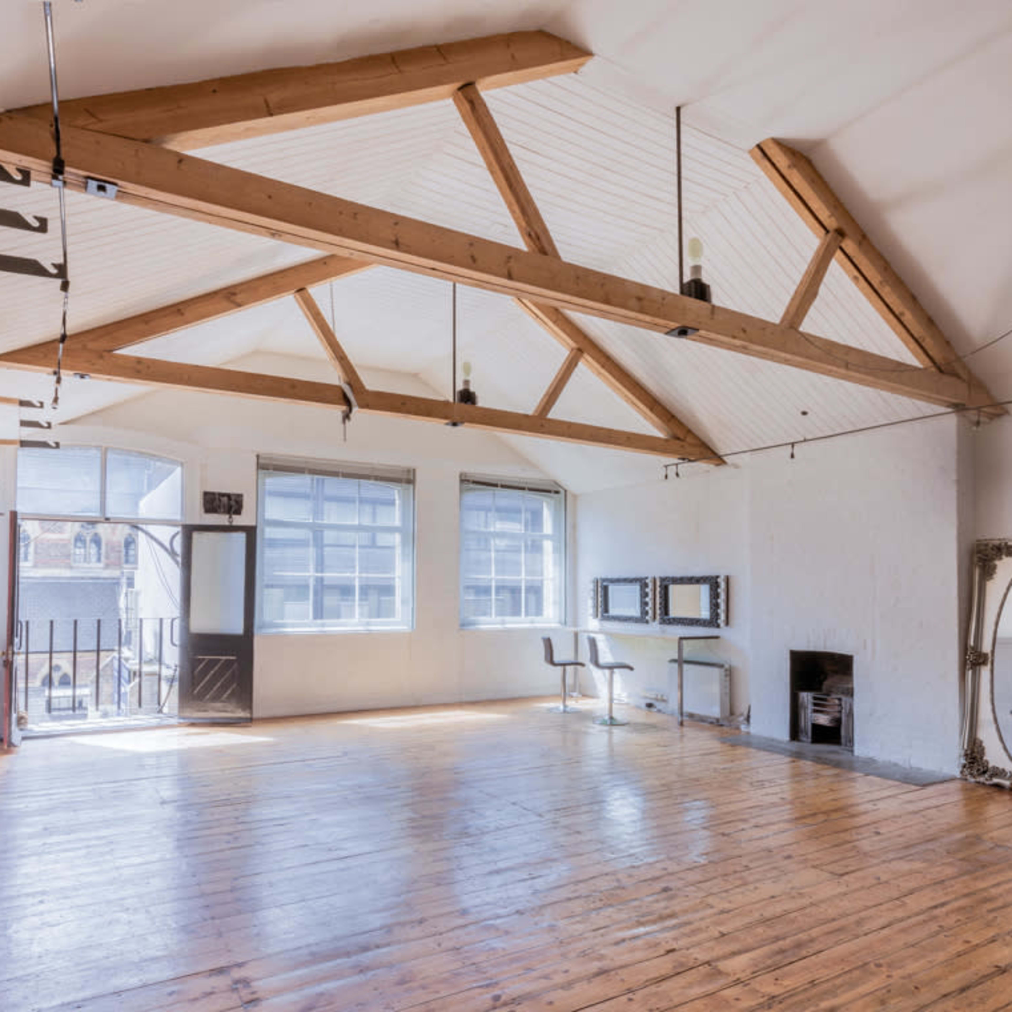 A spacious, well-lit room with wooden floorboards, high ceilings supported by exposed beams, and large windows.