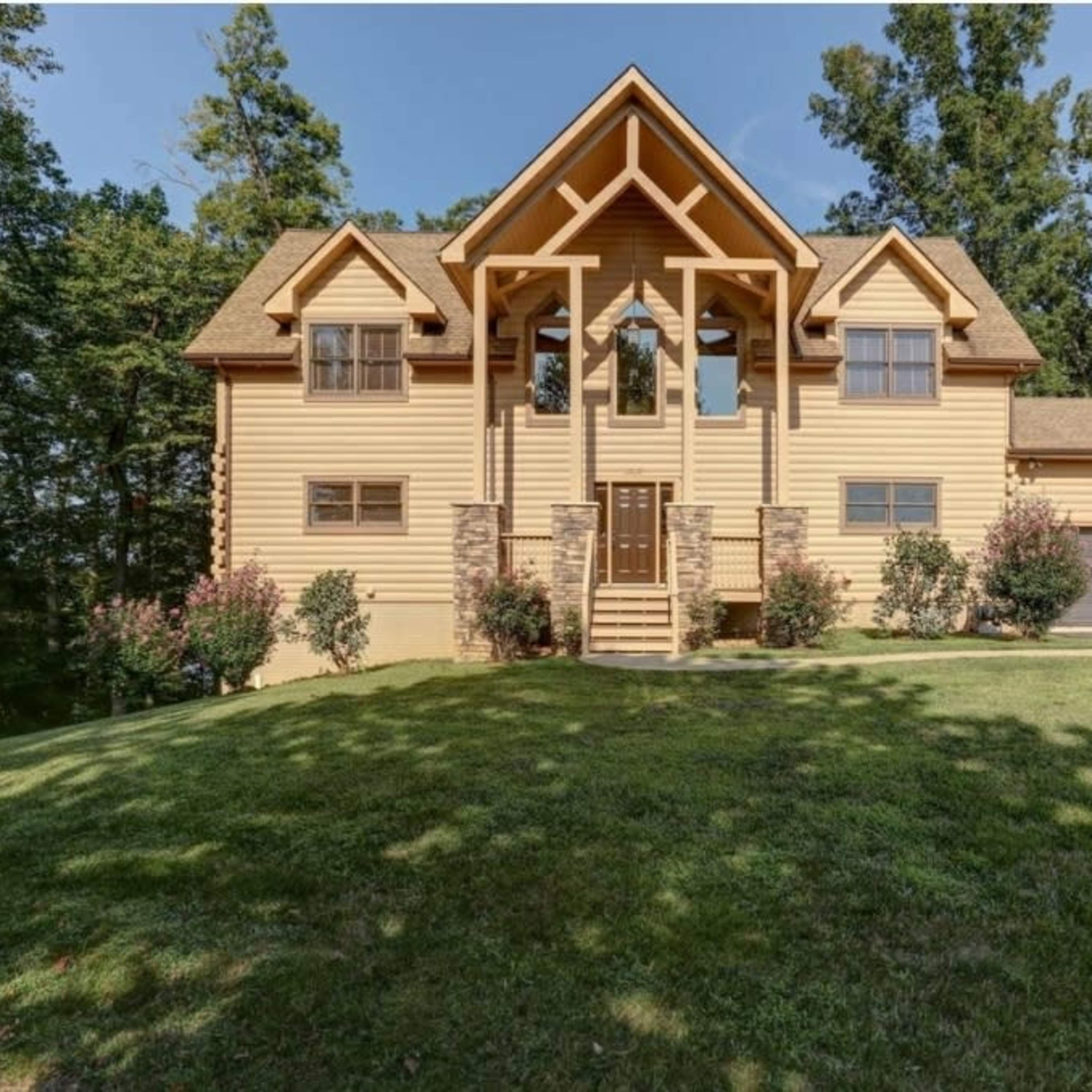 A large, two-story wooden house with a peaked roof, situated on a grassy hill surrounded by trees.