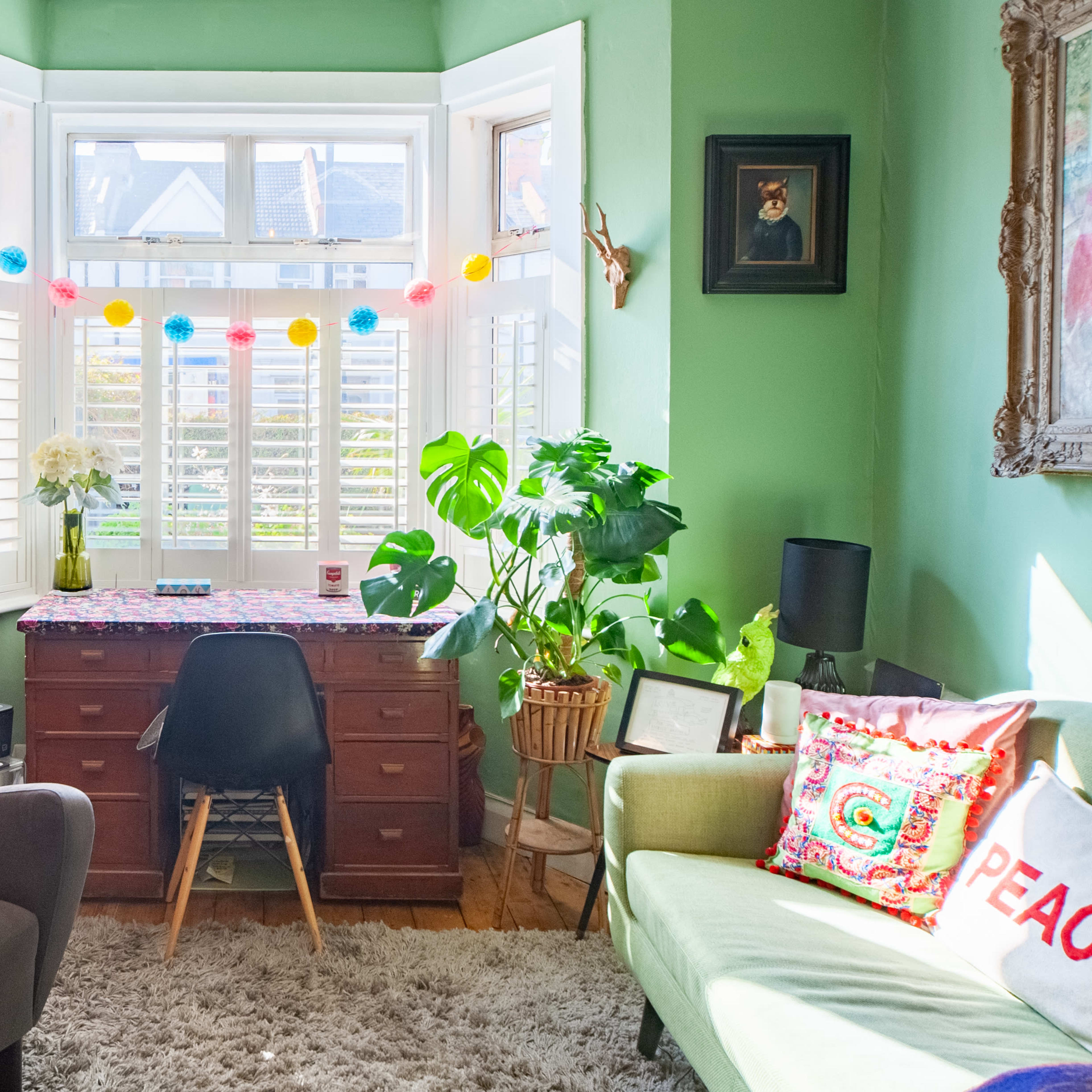 A cozy study features a wooden desk in front of a bay window, surrounded by plants, bookshelves, and comfortable seating.