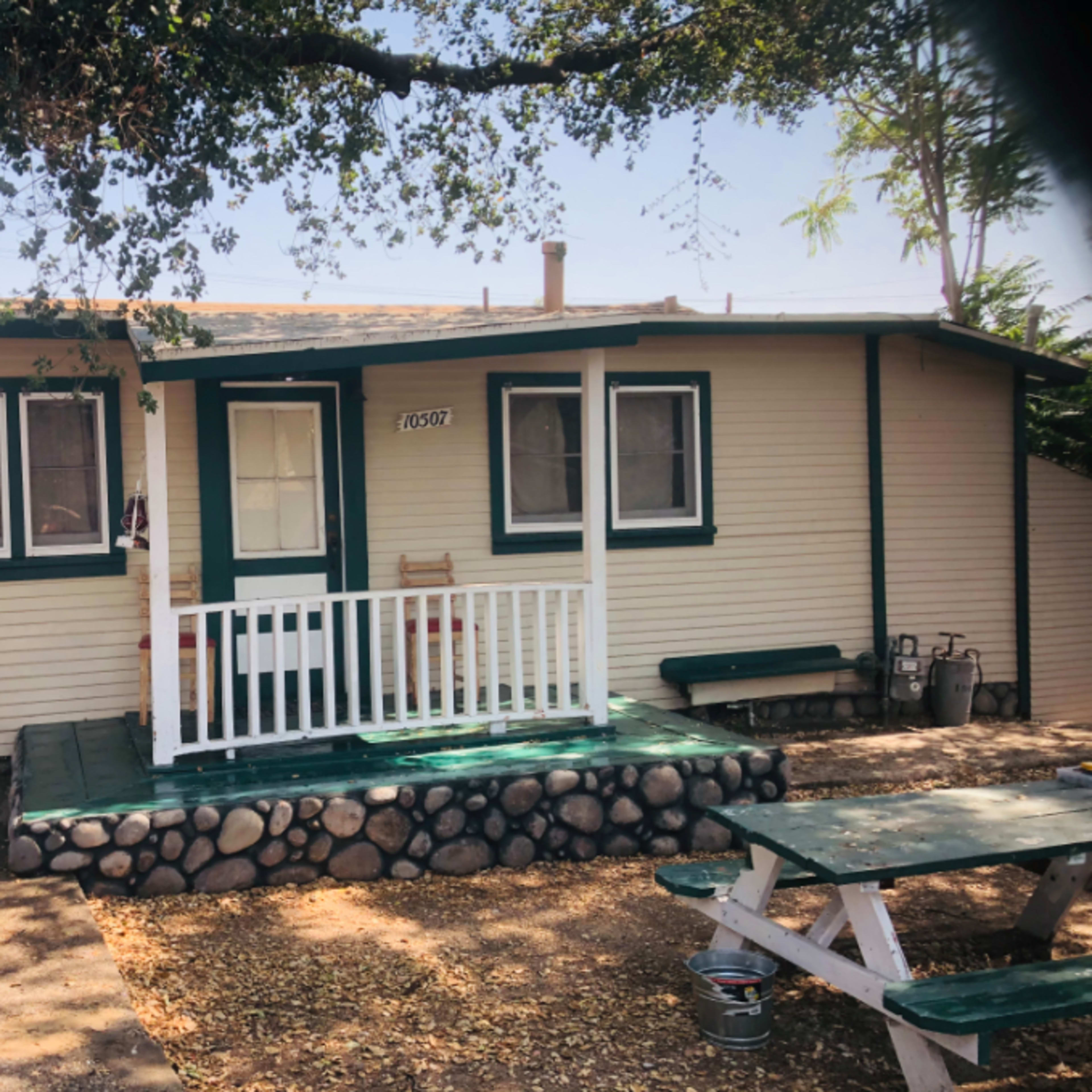 A small, single-story house with a porch and green trim, surrounded by a gravel yard and a picnic table.