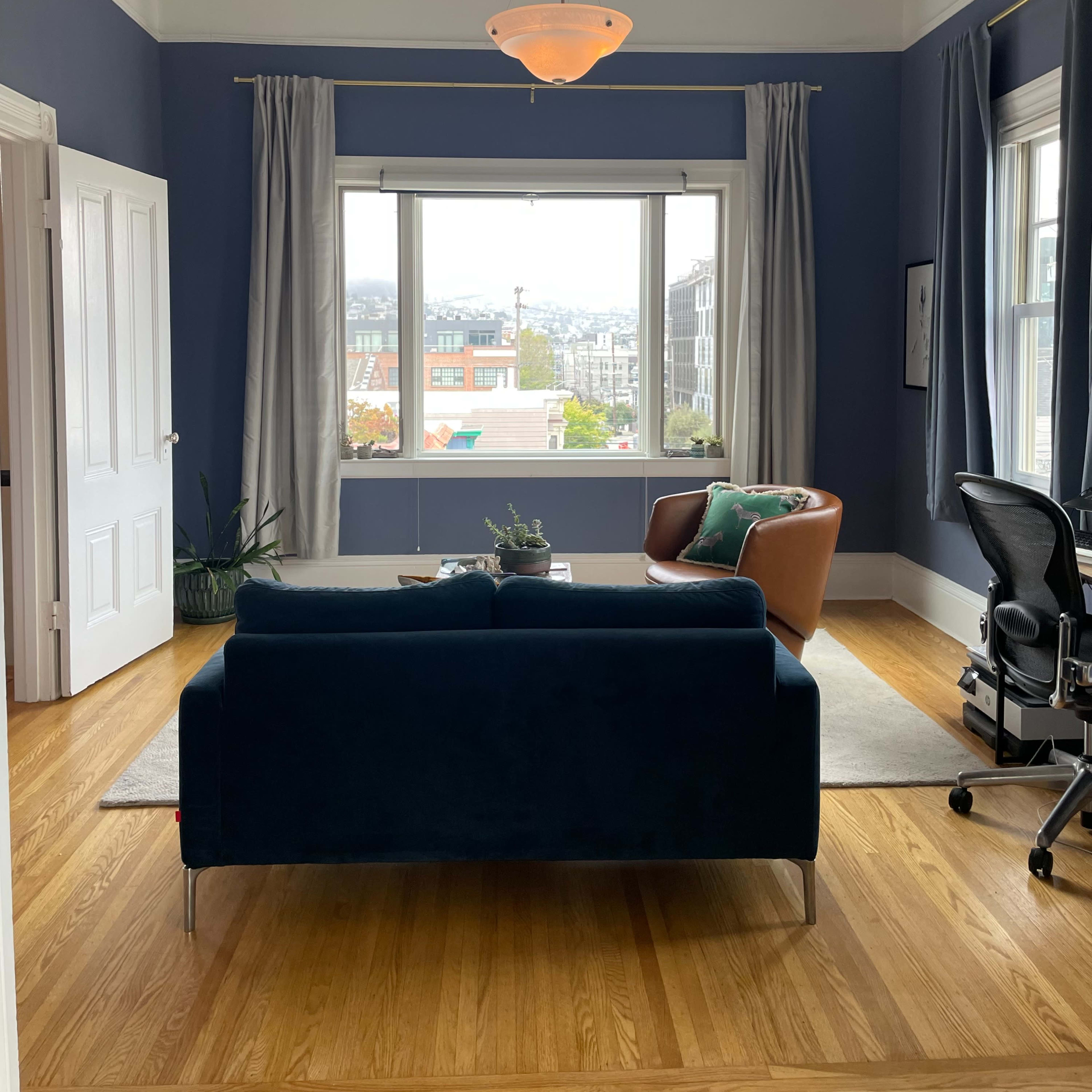 A styled living room featuring a blue couch, a brown armchair, and a view of the city through large windows.