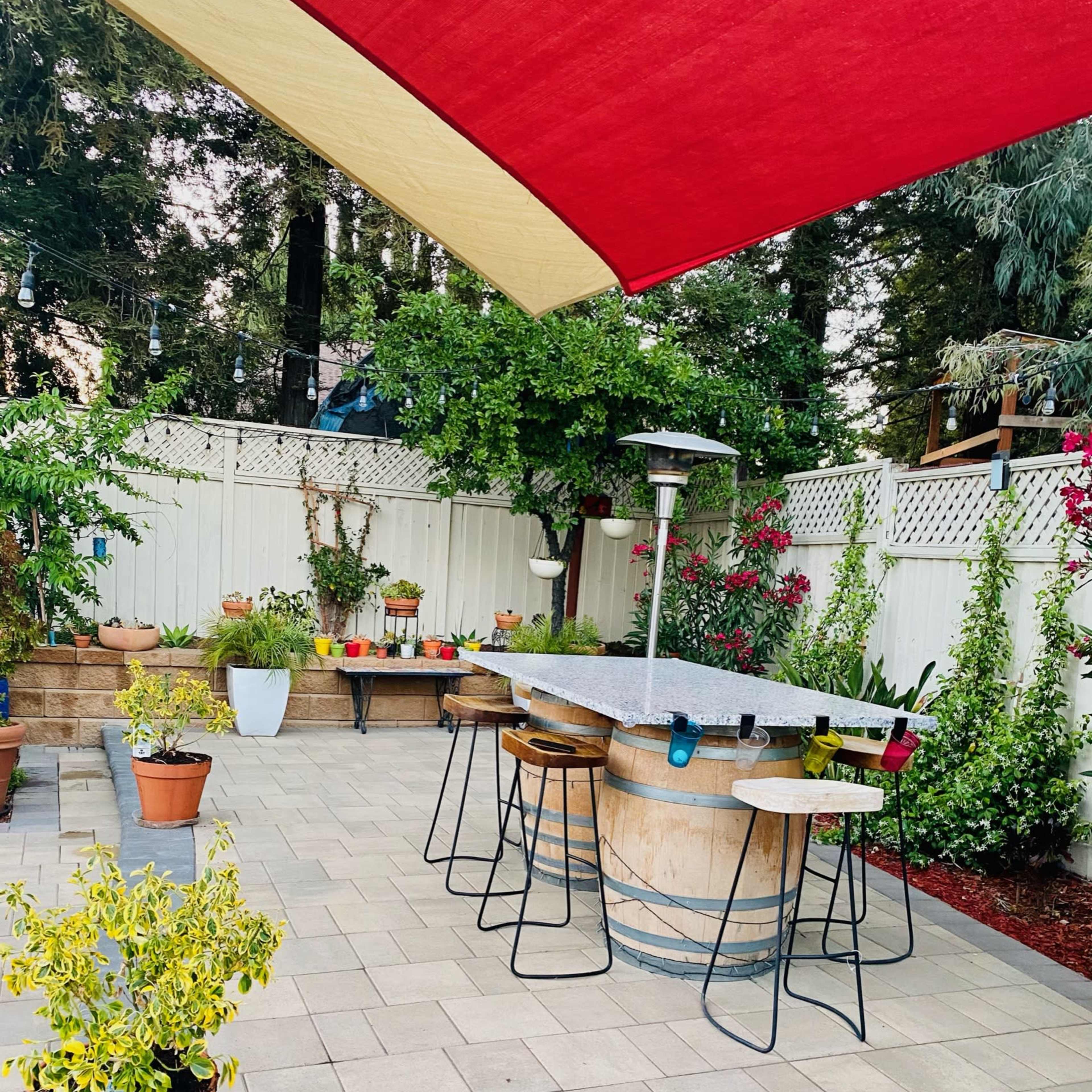 A patio area features a circular stone table with bar stools, surrounded by potted plants and a shaded awning.