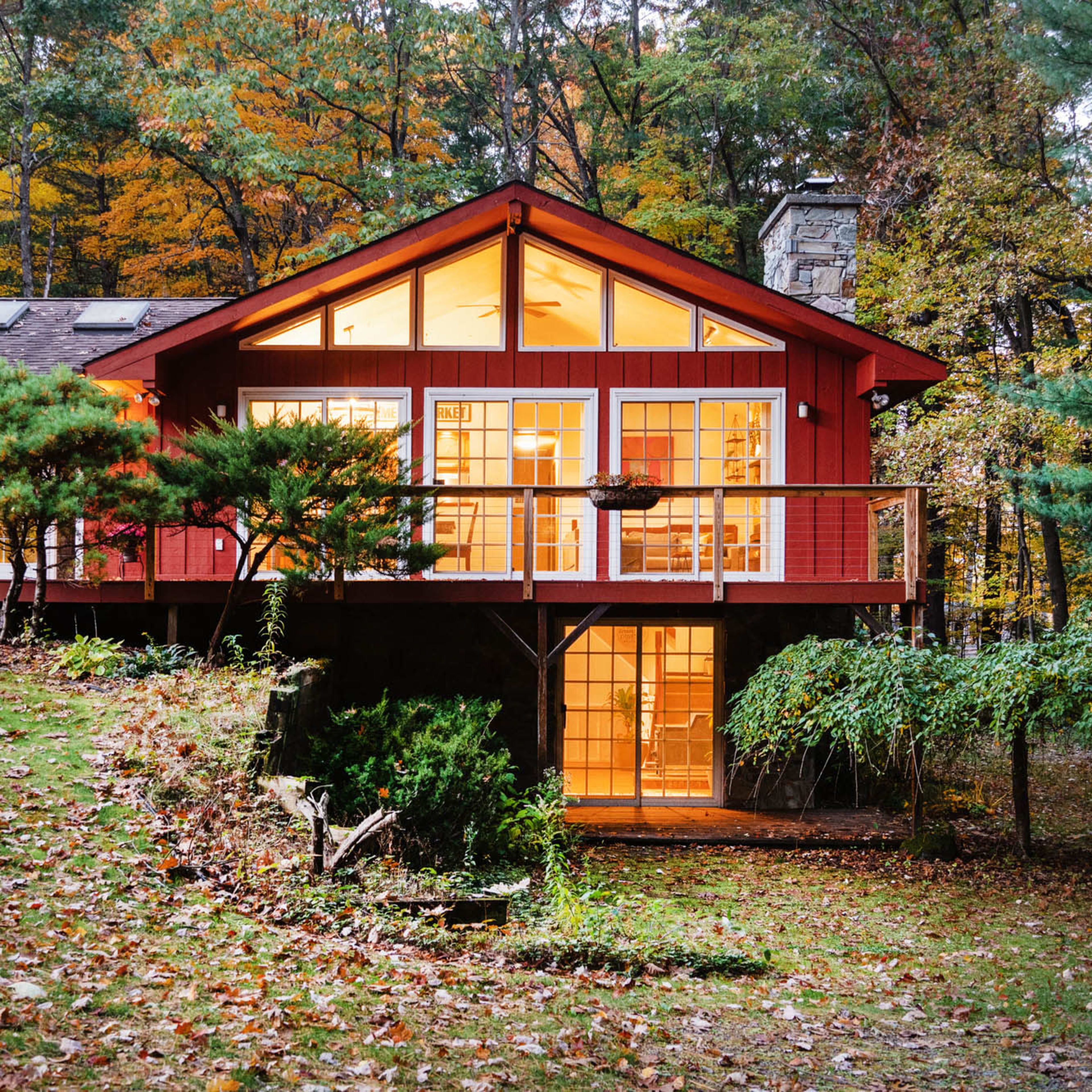 A red cabin with large windows overlooks a landscaped yard surrounded by trees displaying autumn foliage.