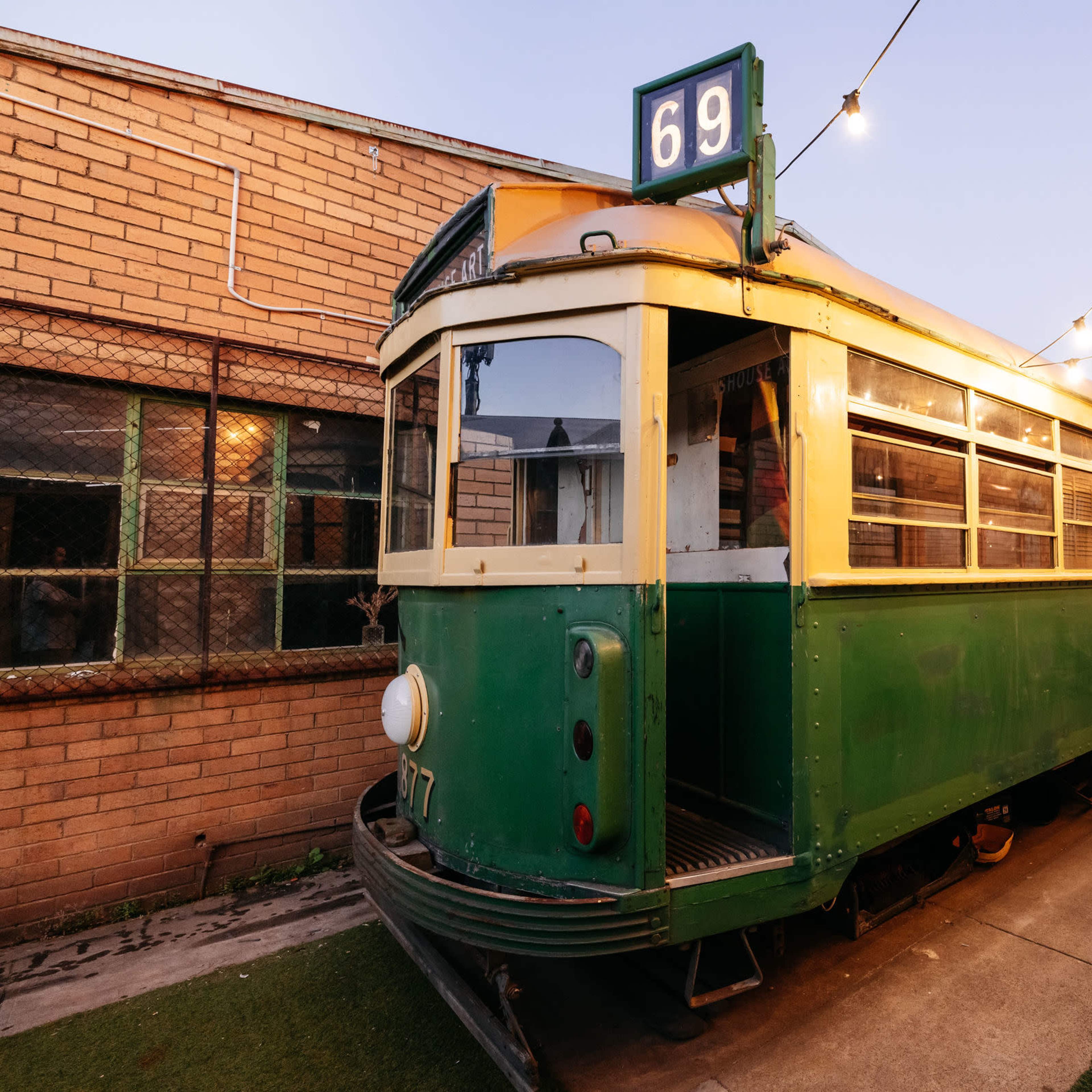 A vintage green tram is parked along a pathway illuminated by string lights at dusk.