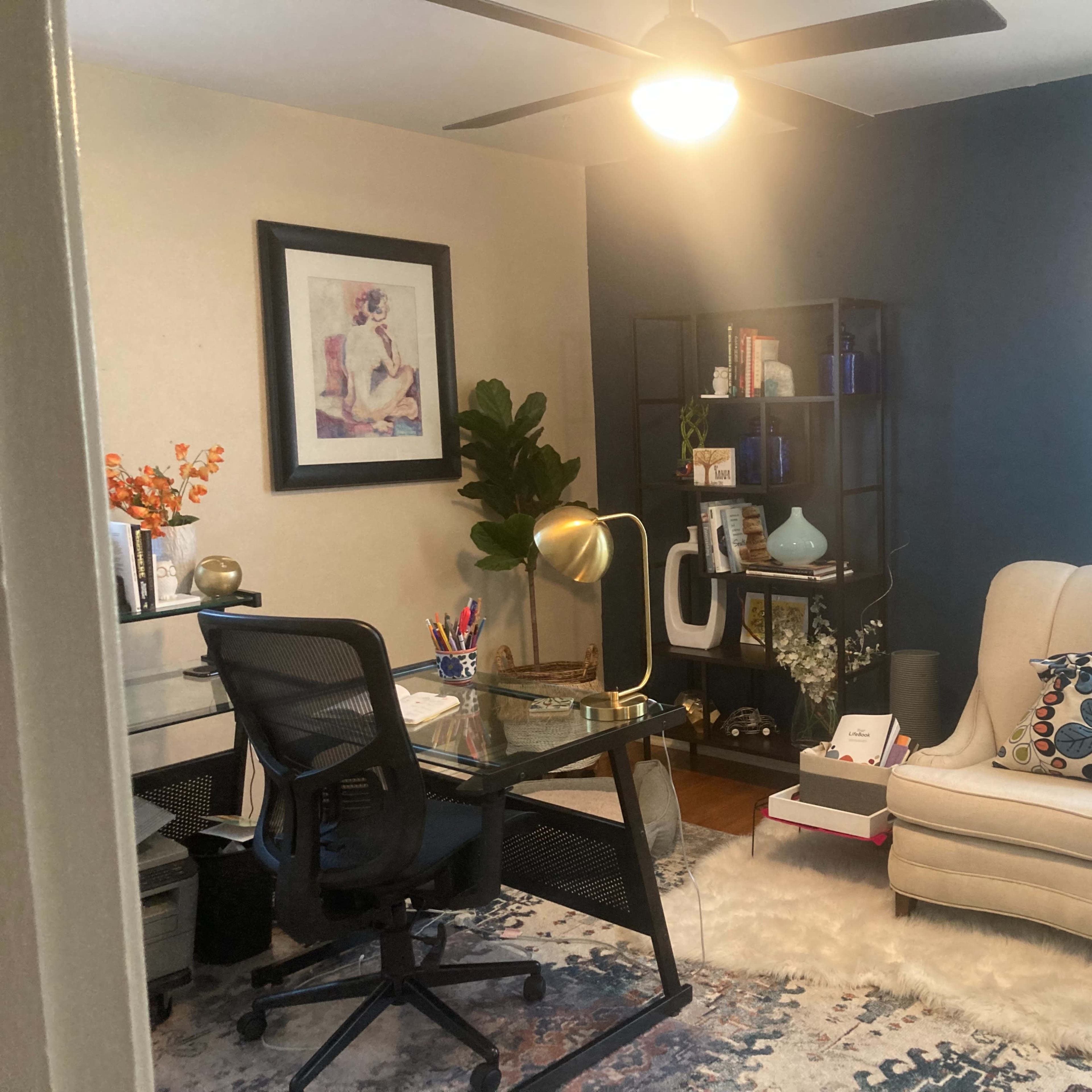 A home office with a desk and chair, a light-colored armchair, shelves filled with various items, and a large rug on the floor.