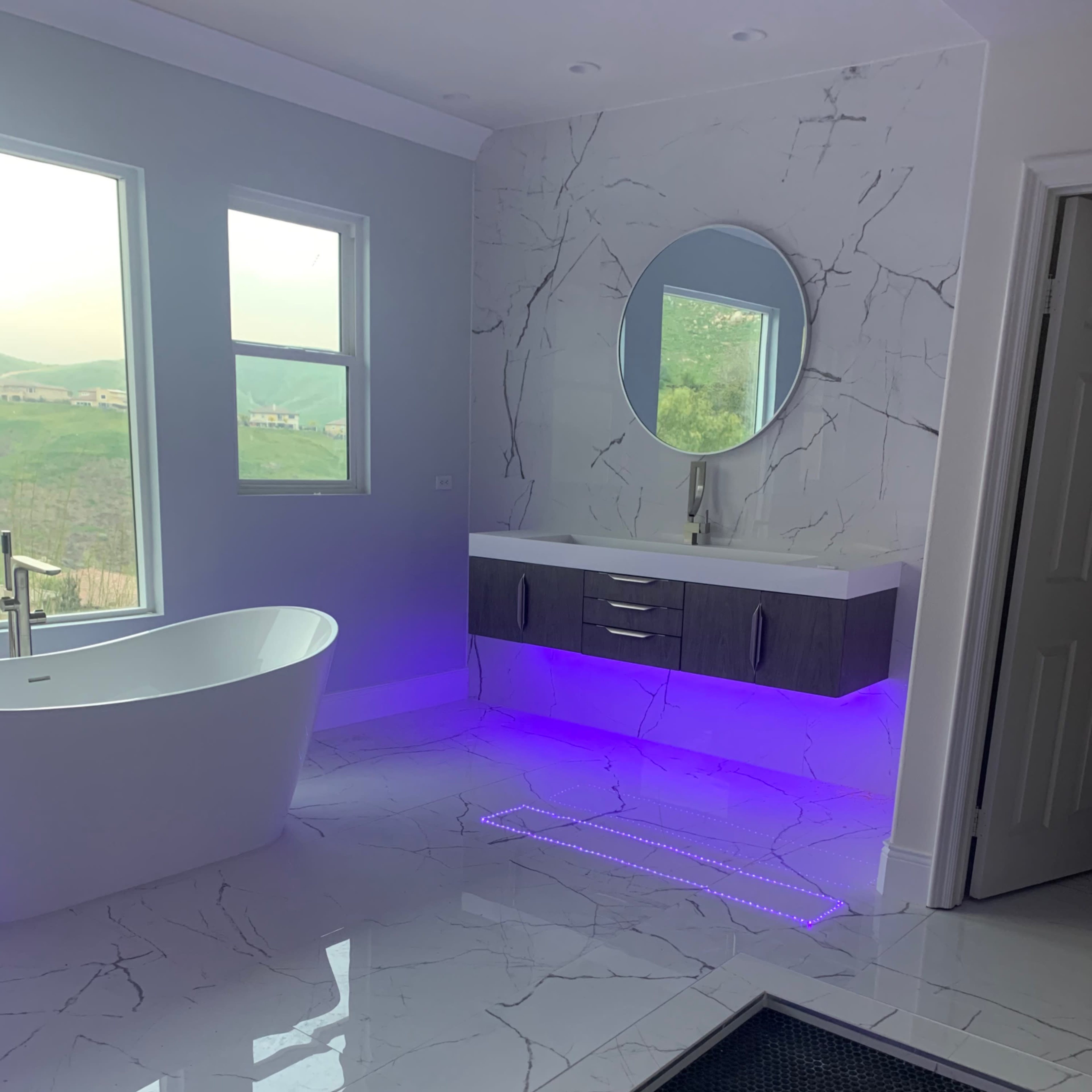 A modern bathroom featuring a freestanding bathtub, a wall-mounted mirror, and a vanity with purple under-lighting.