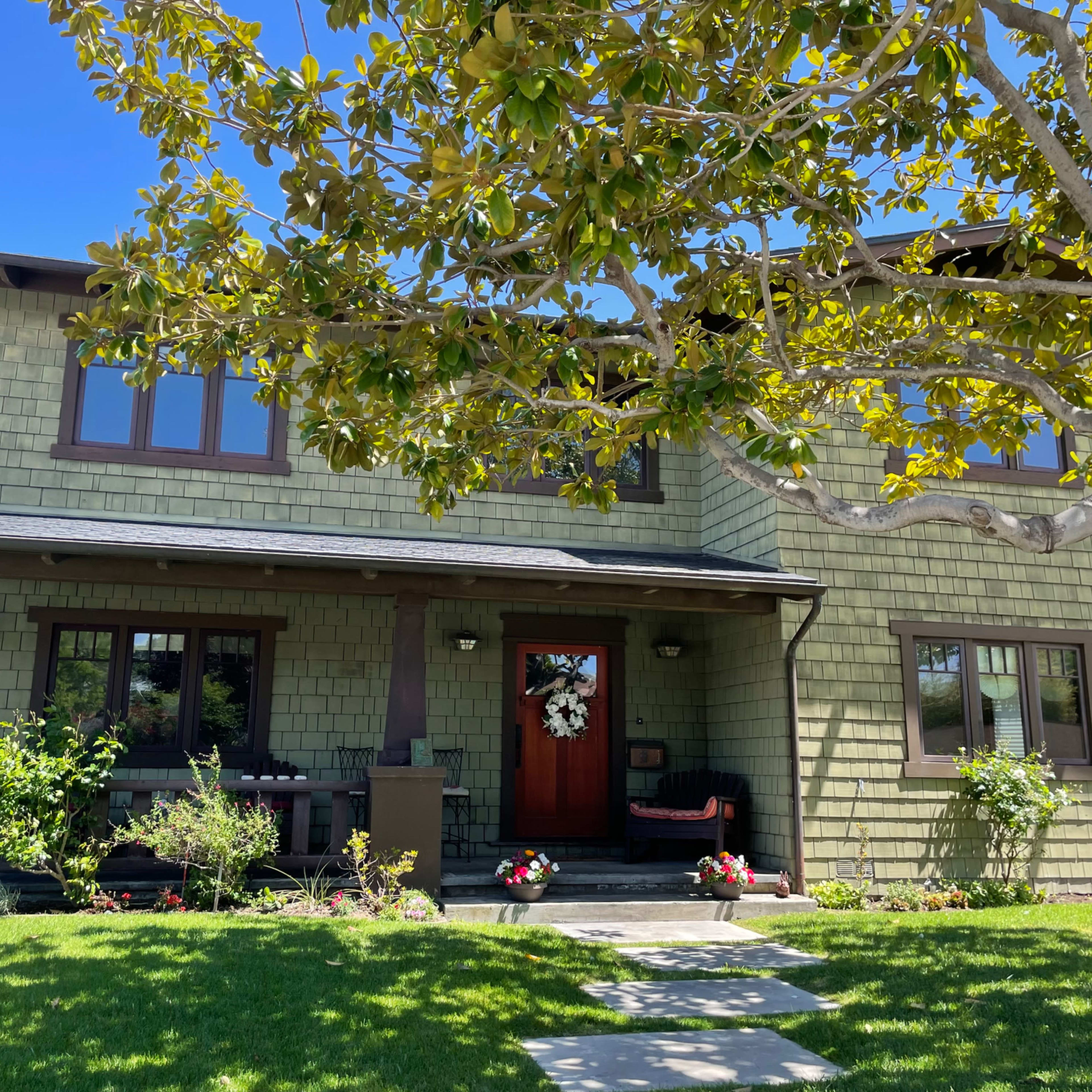 A two-story, green-shingle house with a front porch and a wreath on the door is surrounded by a well-maintained lawn and flowering plants.