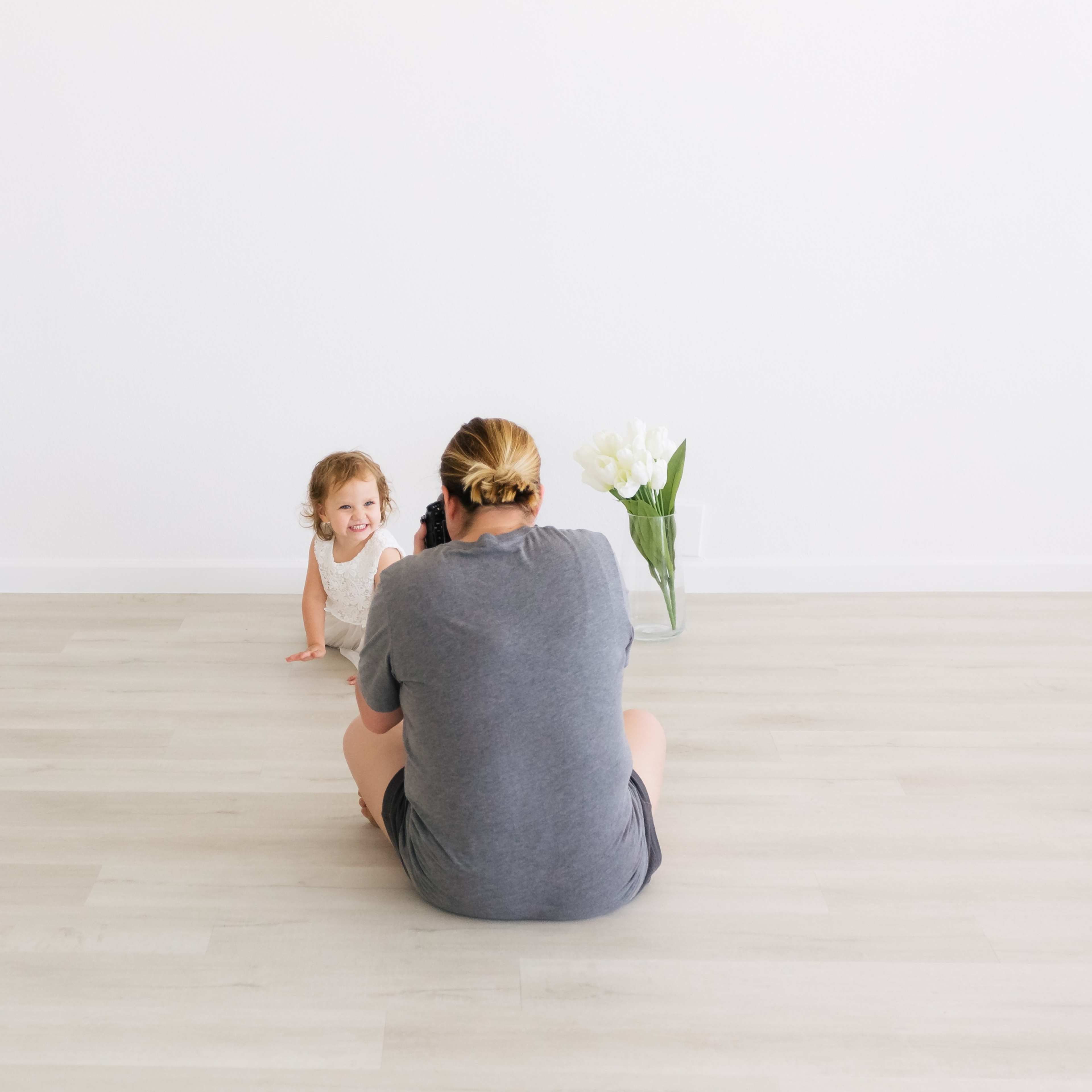 A child crawls toward a person sitting on the floor in a minimalist room with a vase of flowers.