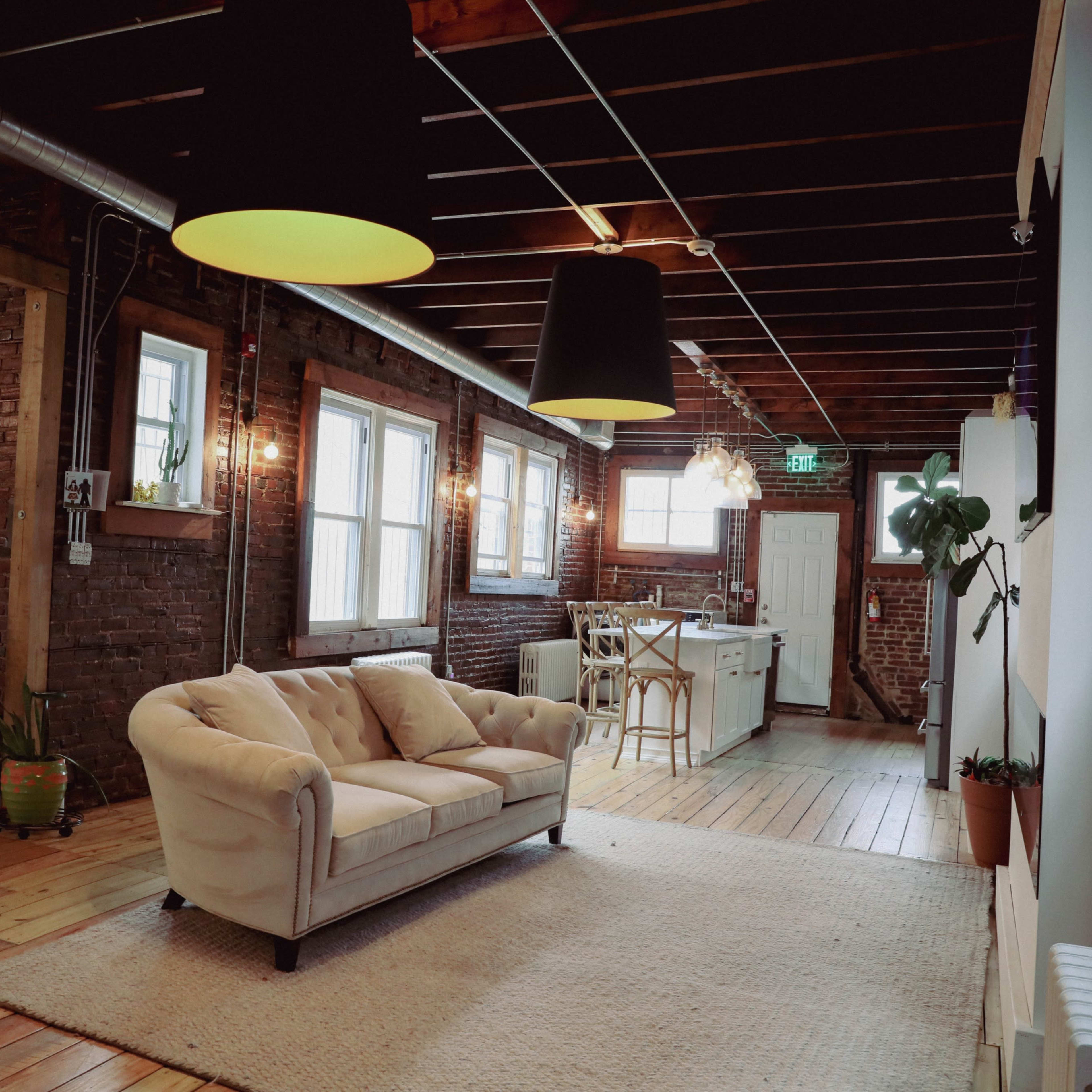 A spacious room with exposed brick walls, featuring a cream-colored couch on a light rug, with large windows and a kitchen area visible in the background.