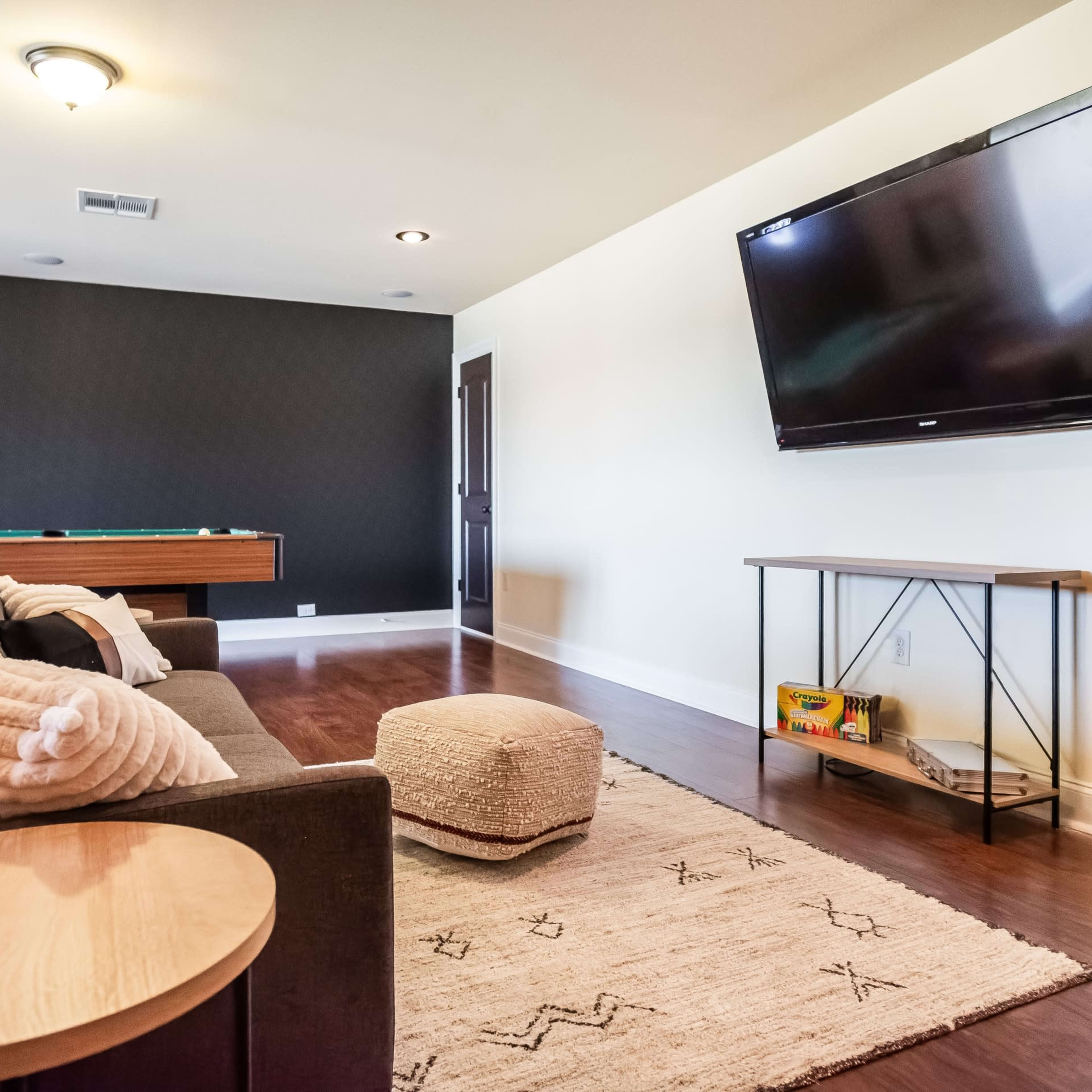 A living room with a television mounted on the wall, a pool table in the background, and a sofa facing a coffee table and a decorative pouf.