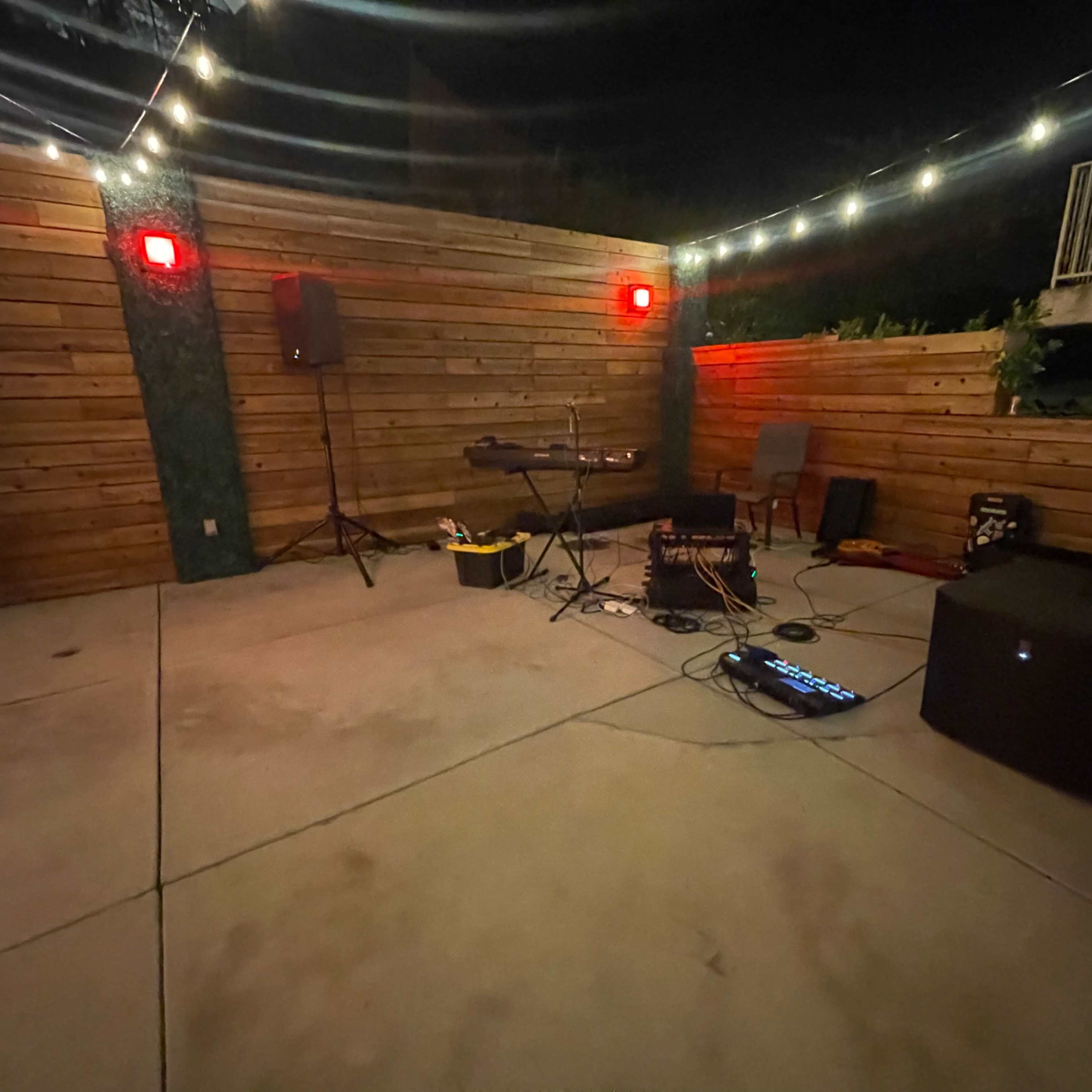 An outdoor patio area at night, featuring wooden walls, string lights, and musical equipment set up on a concrete floor.