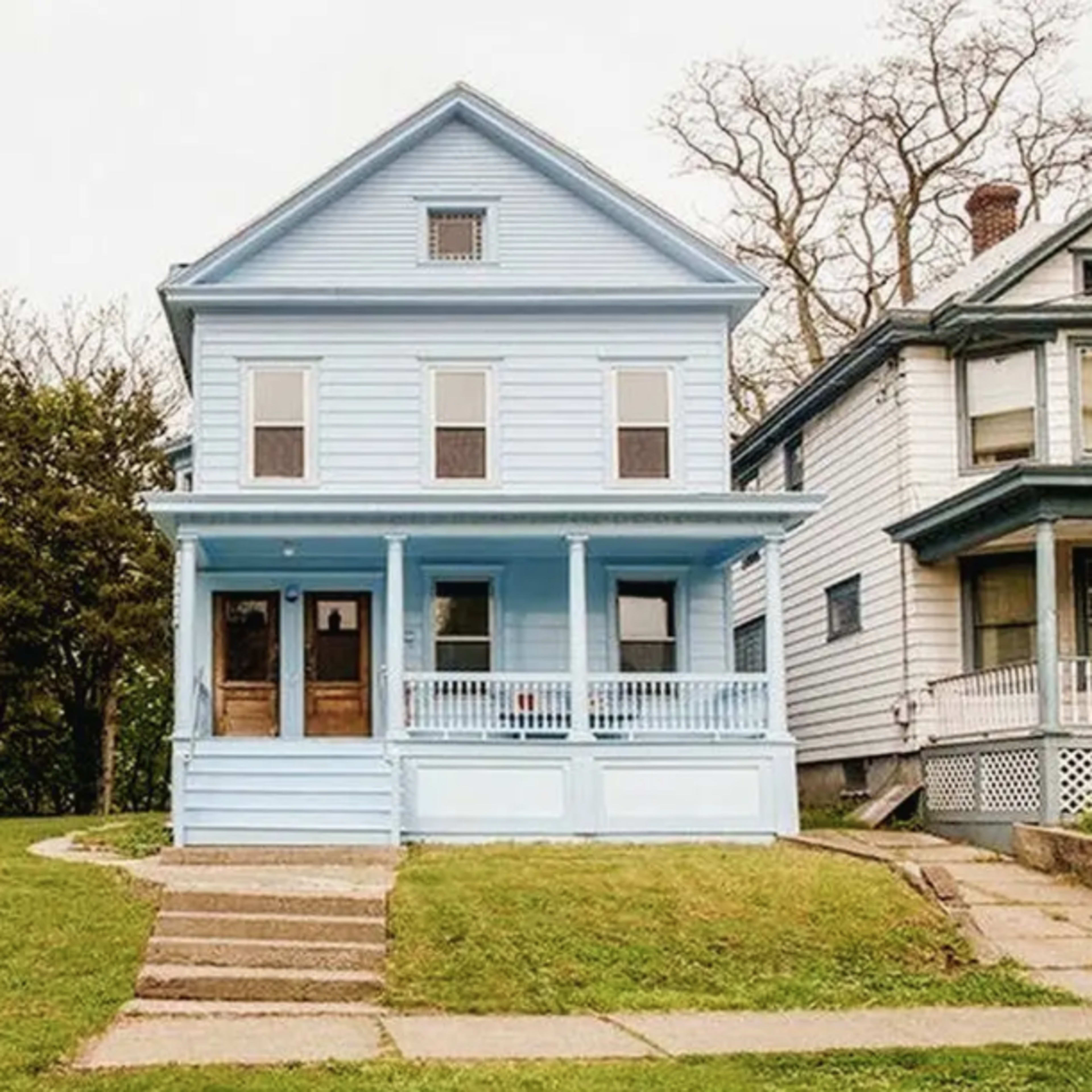 A blue two-story house with a front porch and a grassy yard, situated next to another similar house.