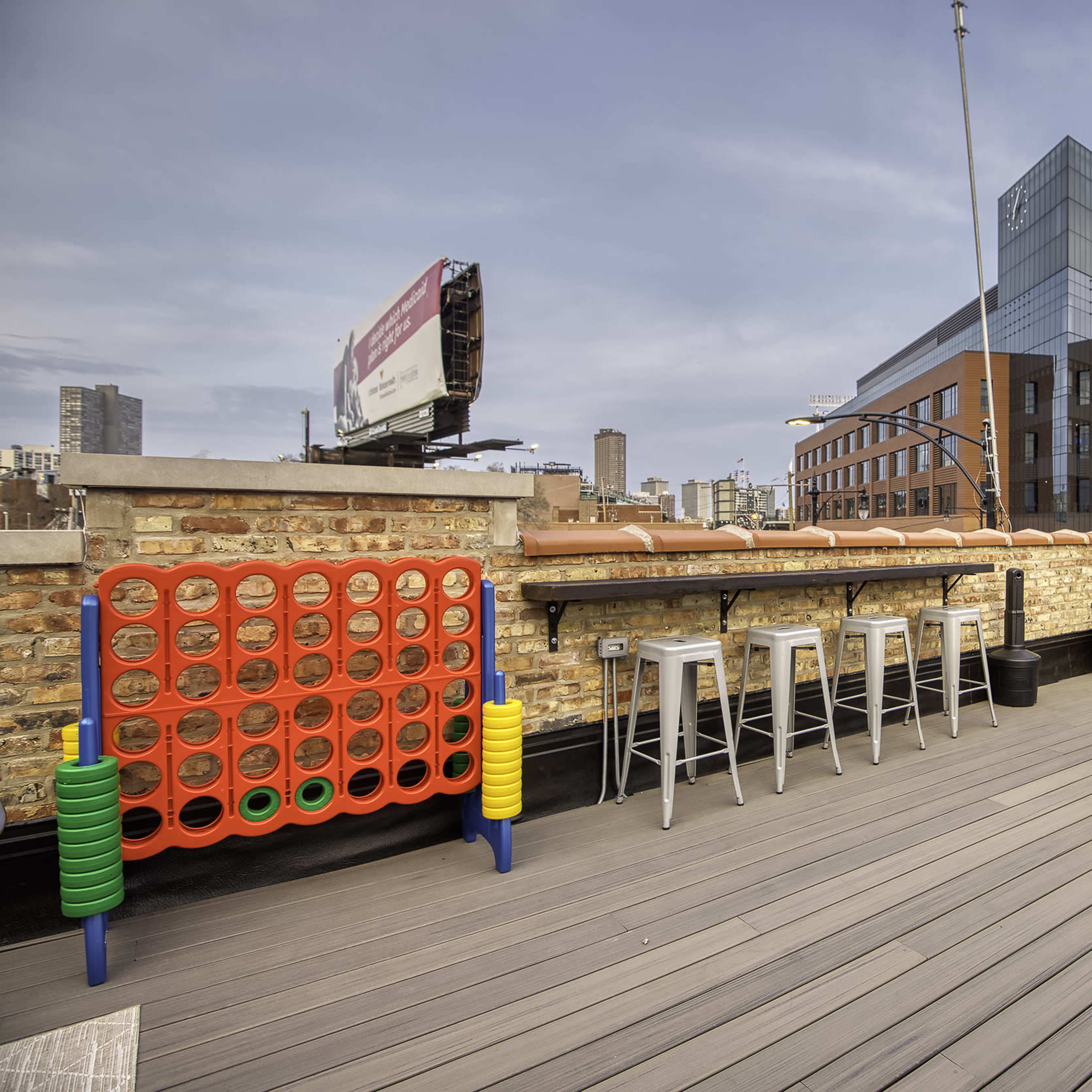 A rooftop terrace featuring a giant Connect Four game, colorful lawn chairs, and a view of surrounding buildings.