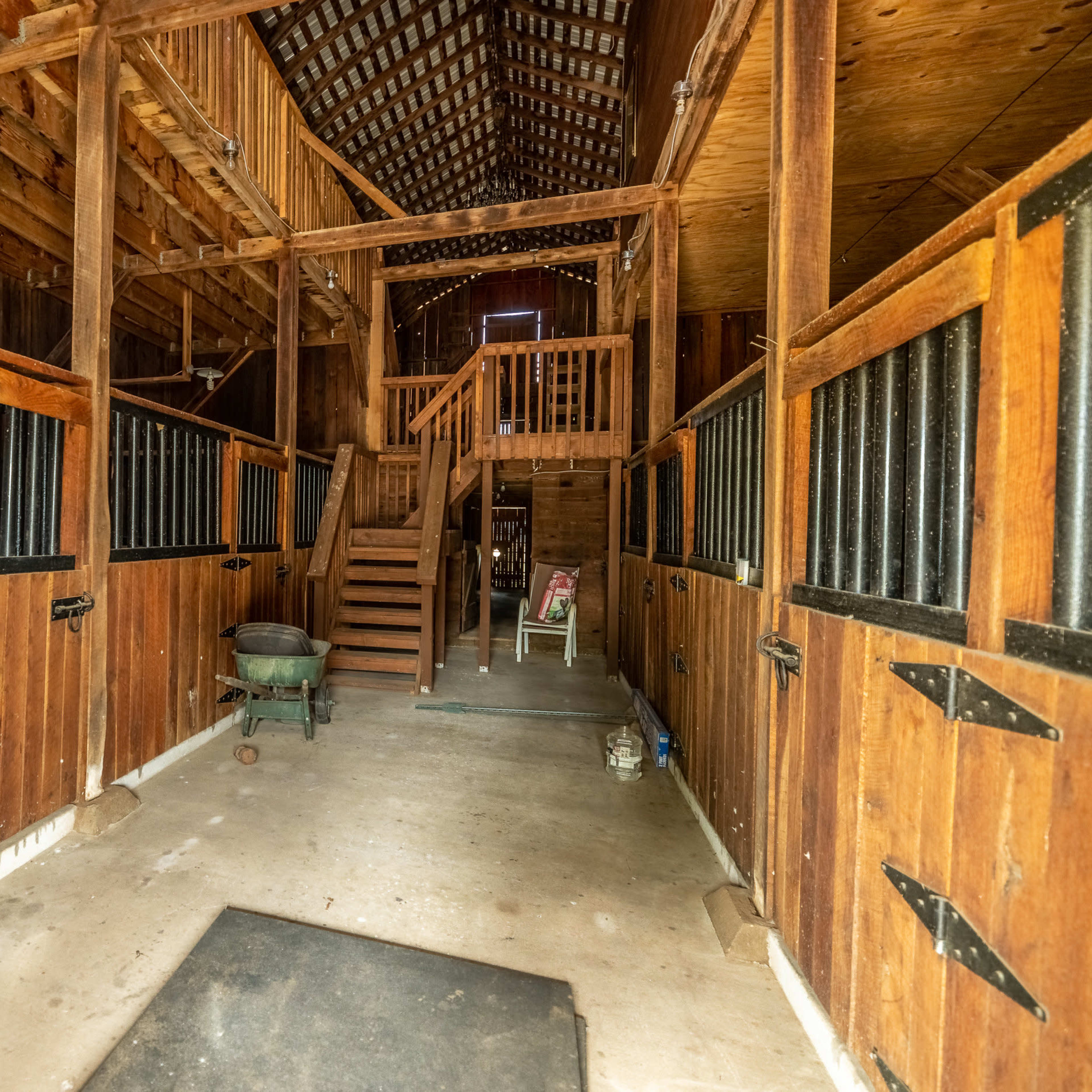 The interior of a rustic barn with wooden stalls, a staircase leading to an upper level, and a concrete floor.