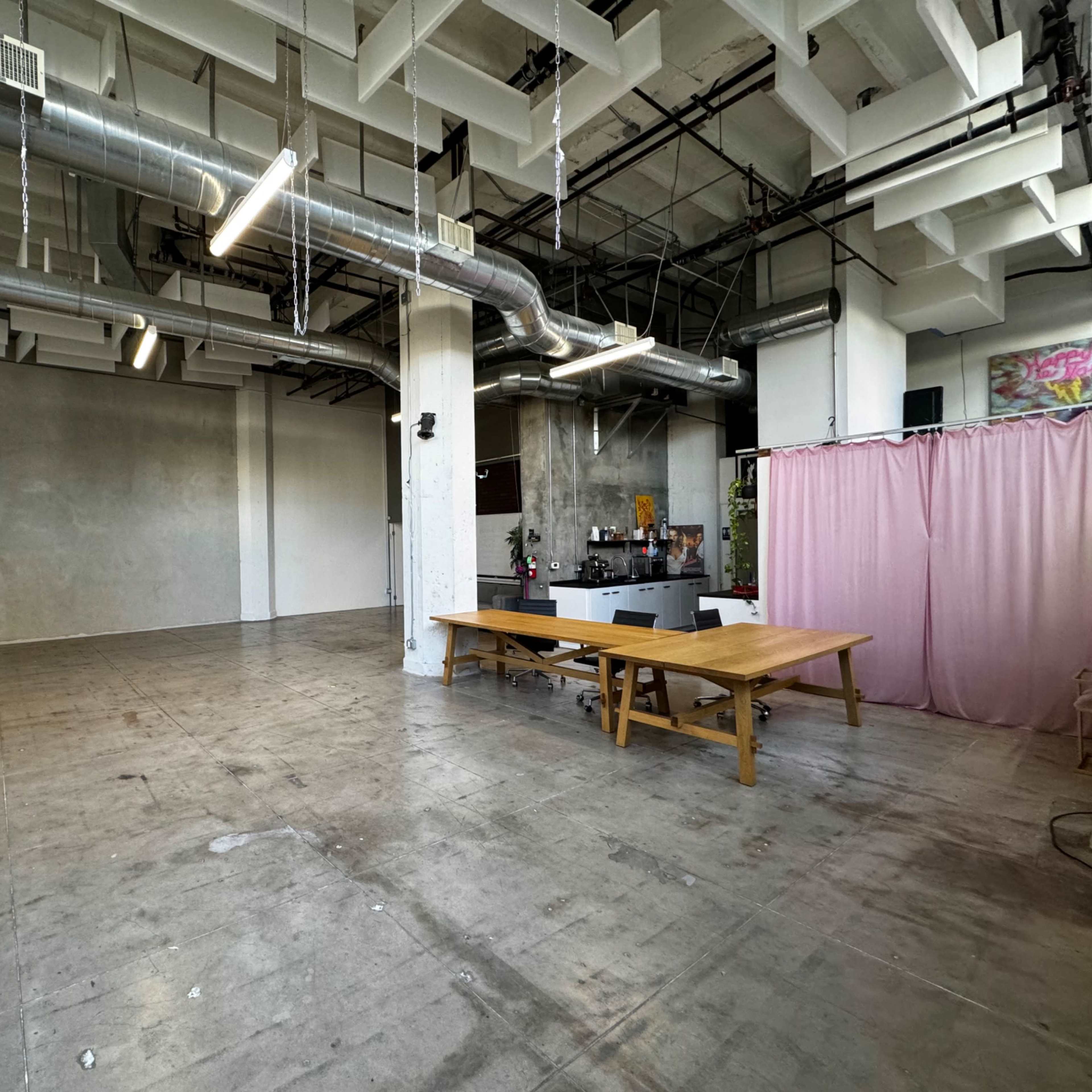 A spacious, industrial-style room with concrete floors, large windows, and a pink curtain separating an area with a kitchen and dining table.