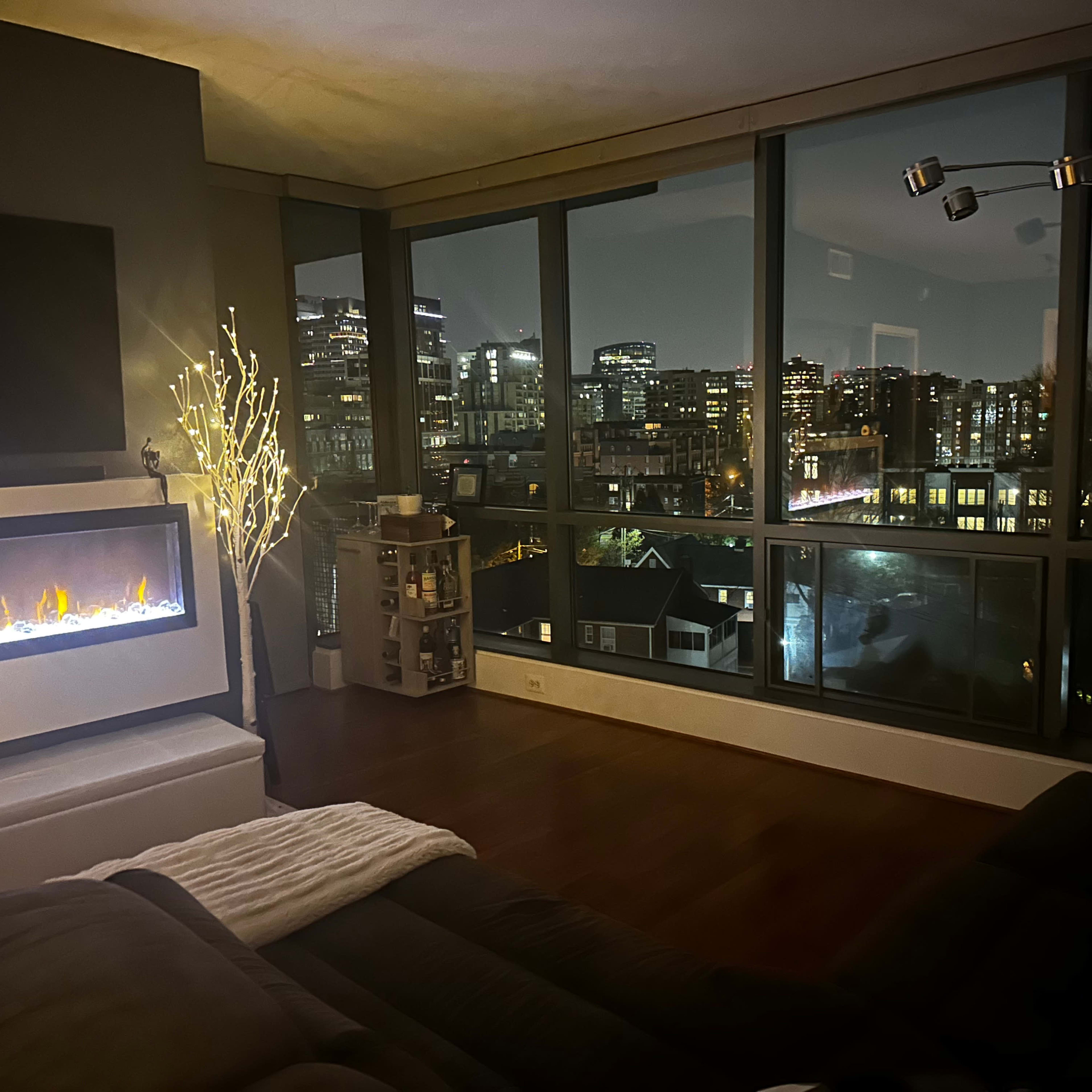 The scene captures a modern living room with large windows overlooking a city skyline at night, featuring a sofa, an LED fireplace, and decorative lighting.