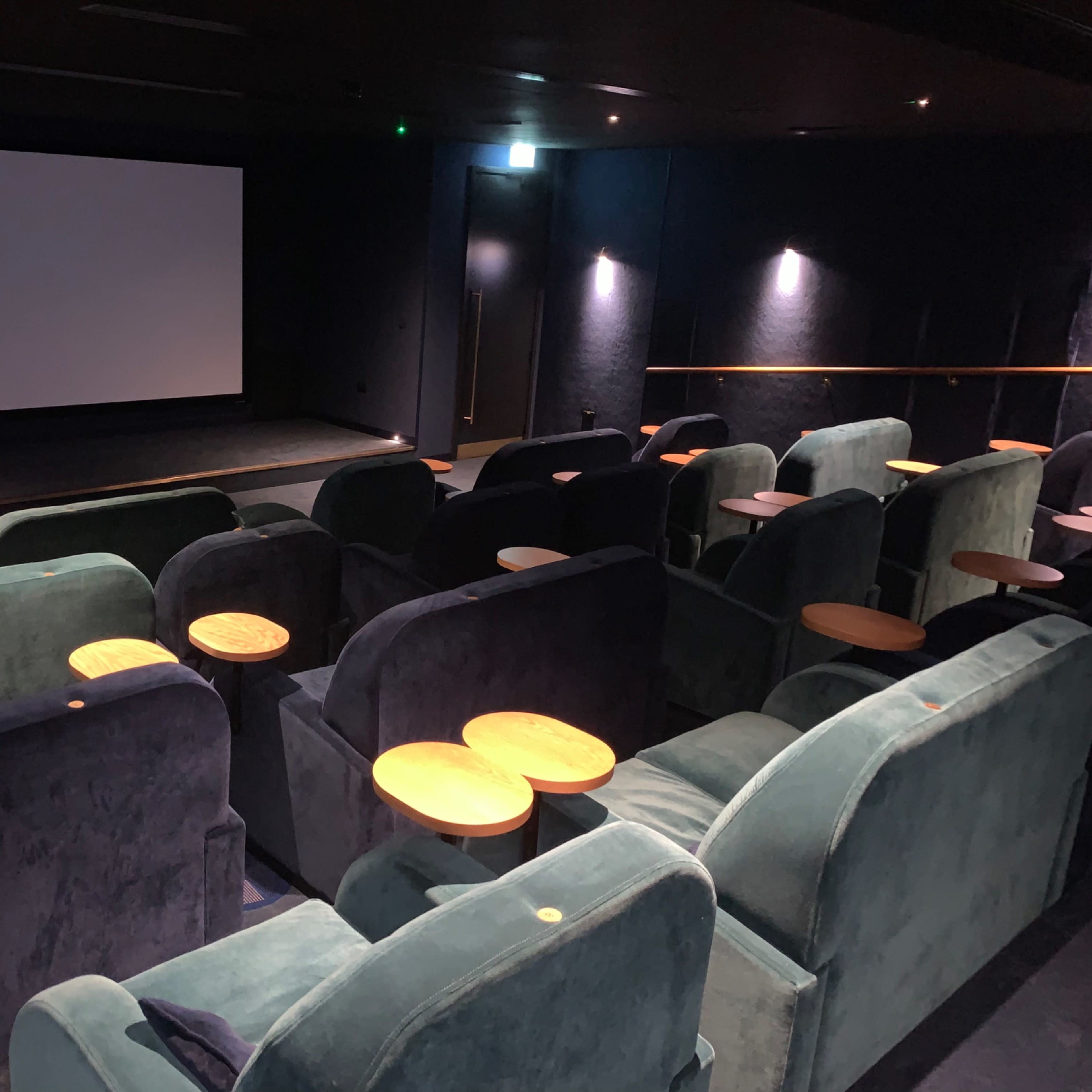 A small, well-arranged movie theater with plush seats and side tables facing a blank screen.