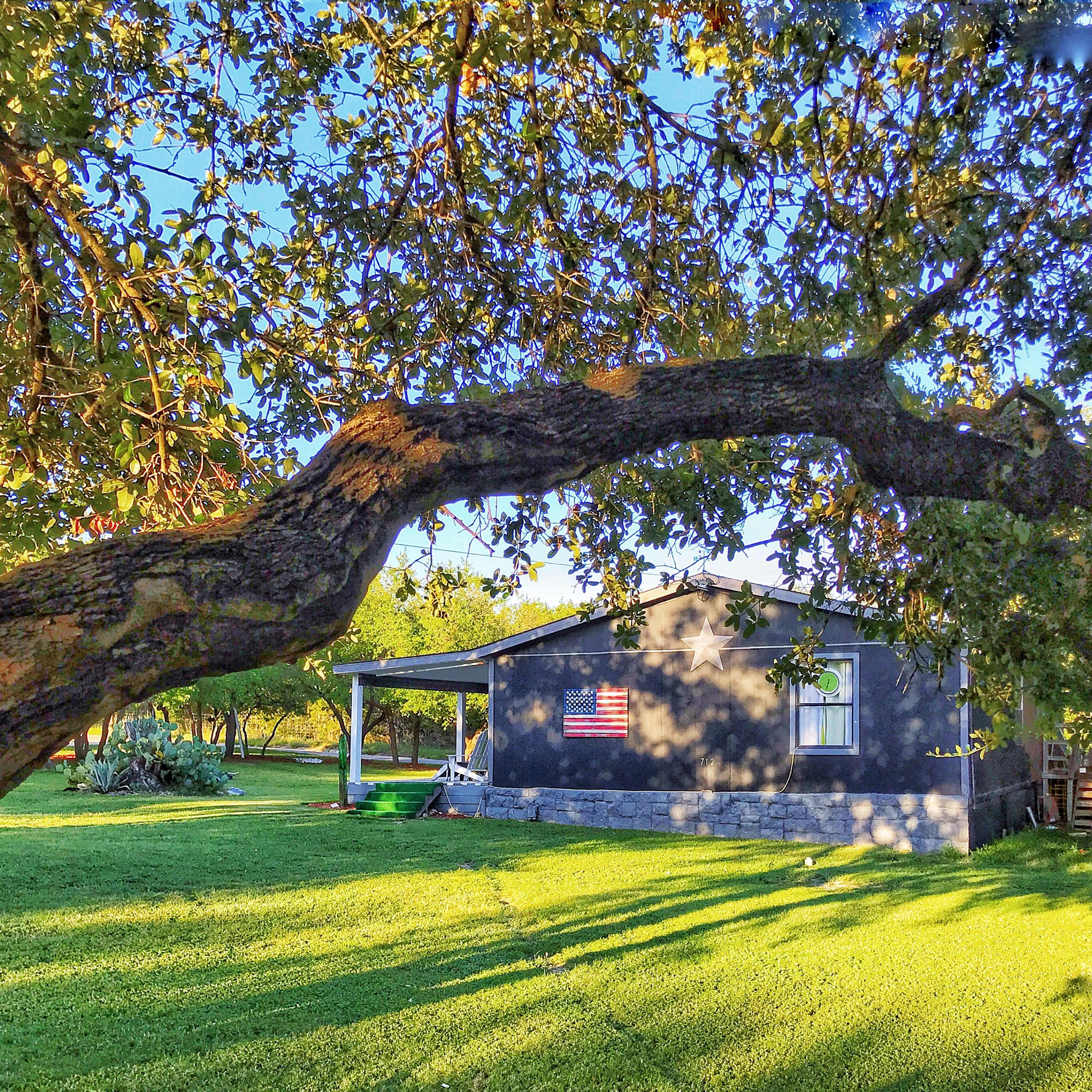 A house with a flag displayed on its side is surrounded by a green yard and shaded by a tree.