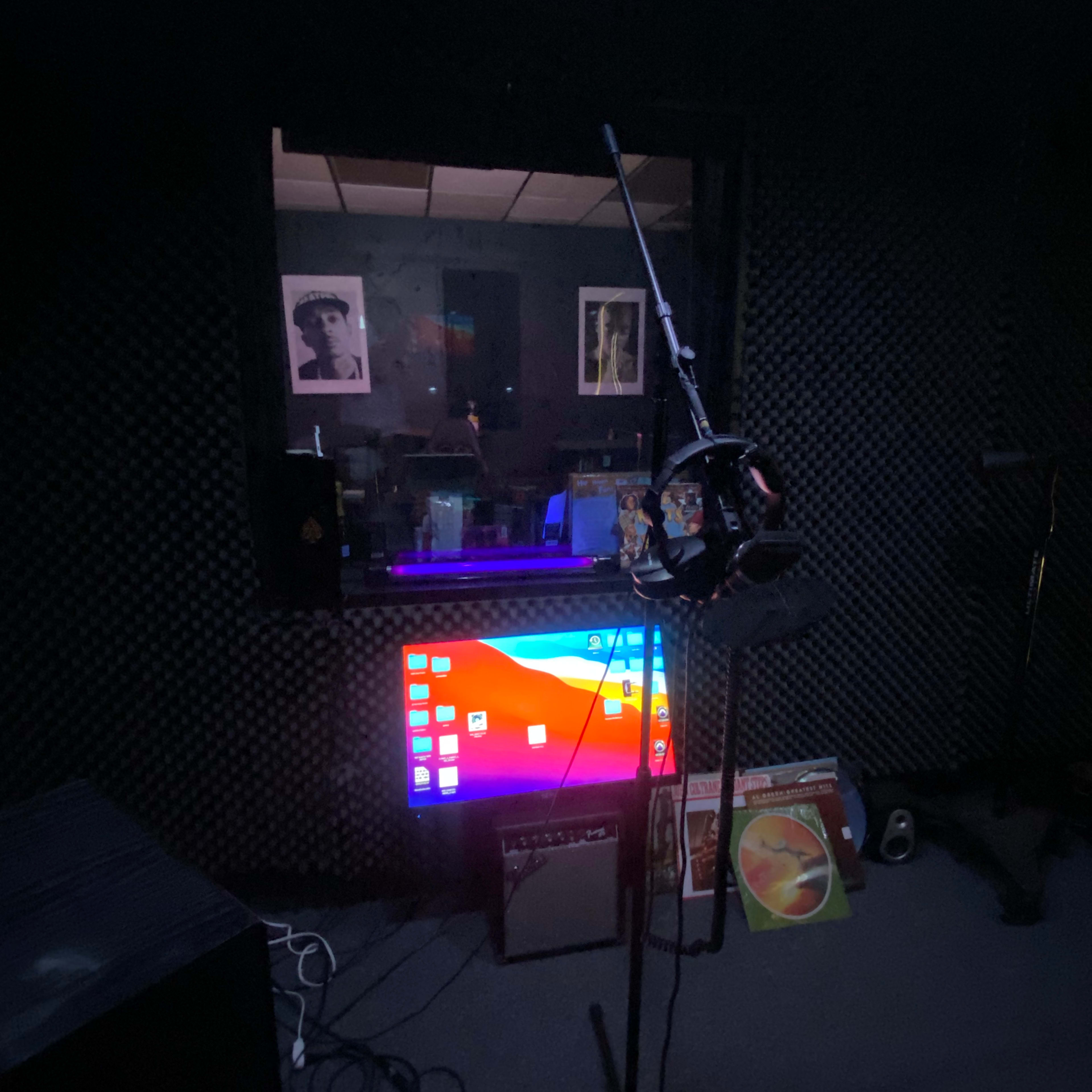 A microphone stand is positioned in front of a soundproofed recording booth, with computer monitors and vinyl records visible through the glass.